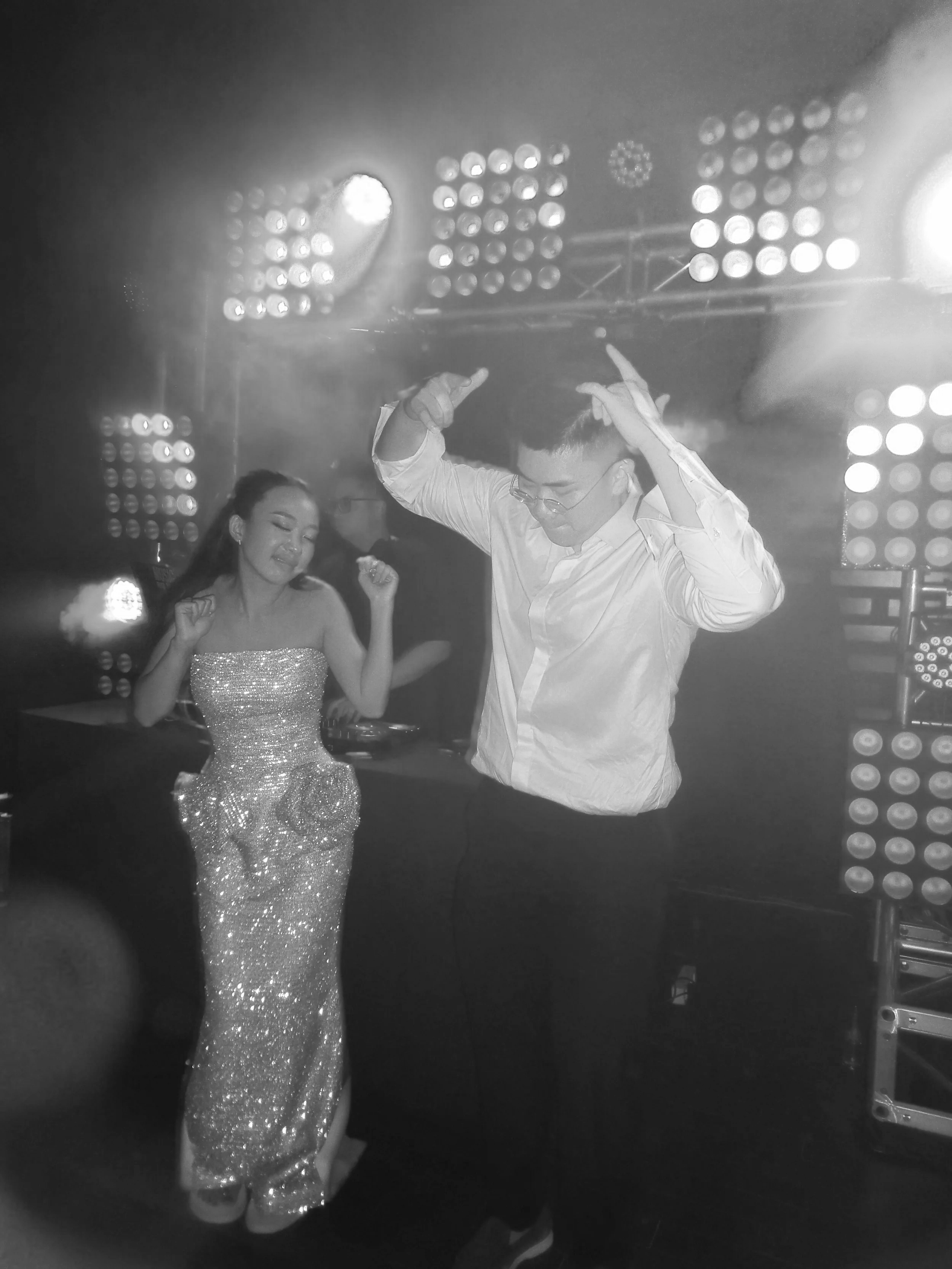 Two people dancing at a nightclub with bright stage lights and DJ equipment in the background, one woman in a sparkling dress and one man in a white shirt.