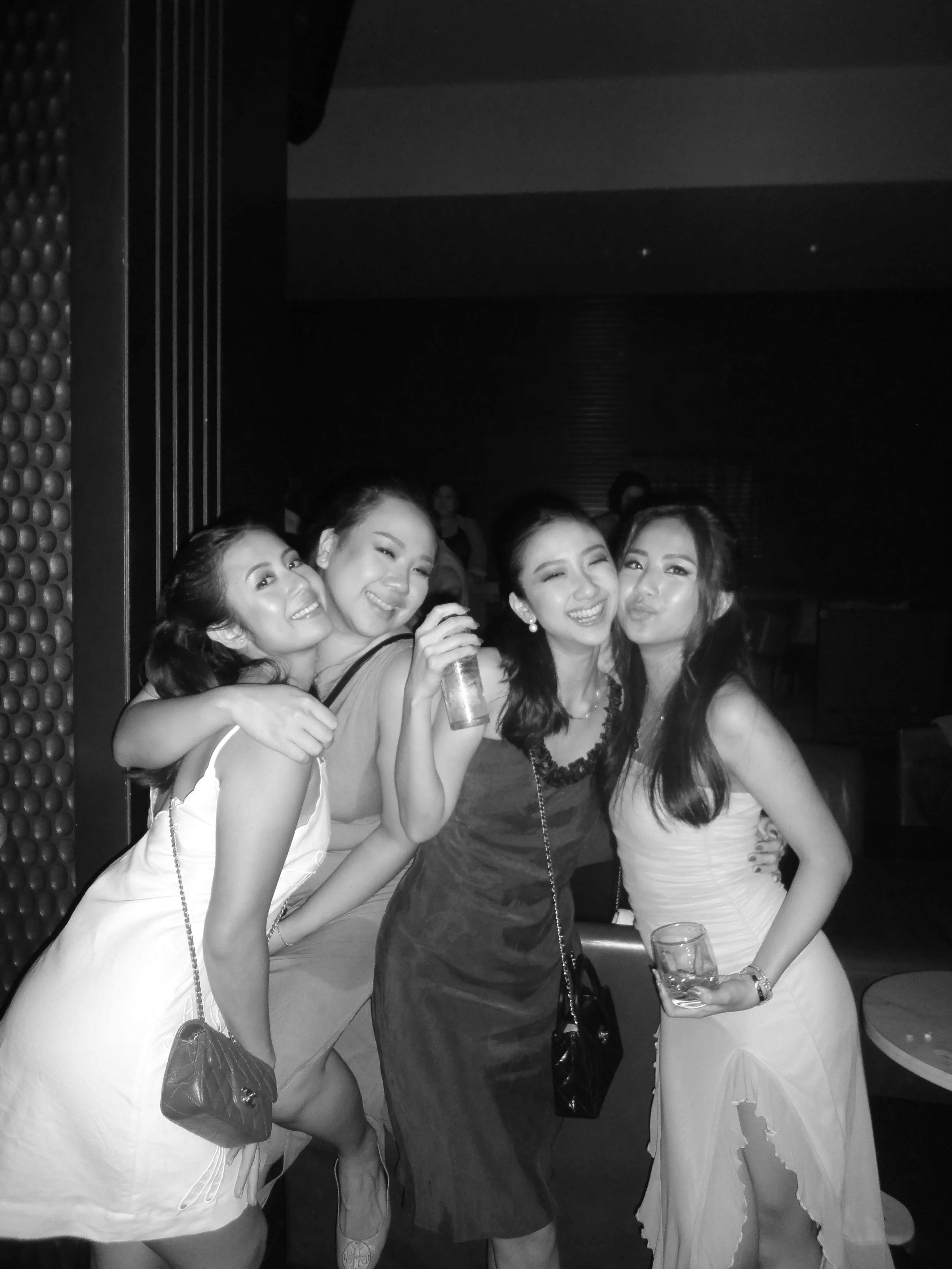 Four women in party dresses smiling and posing together at a social gathering or party, holding drinks.