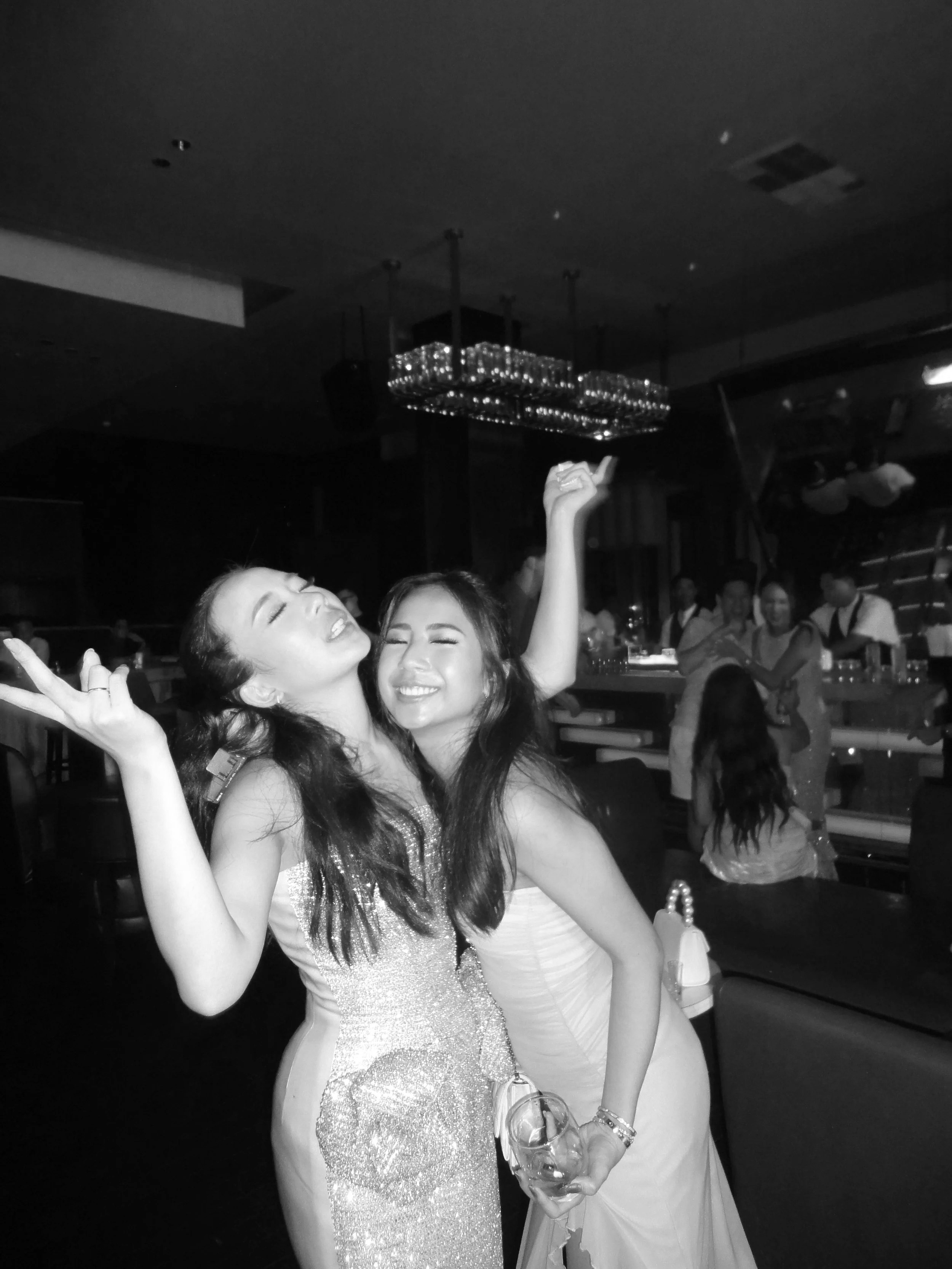 Two women dancing and celebrating at a party or event in a dimly lit venue, with other people in the background.