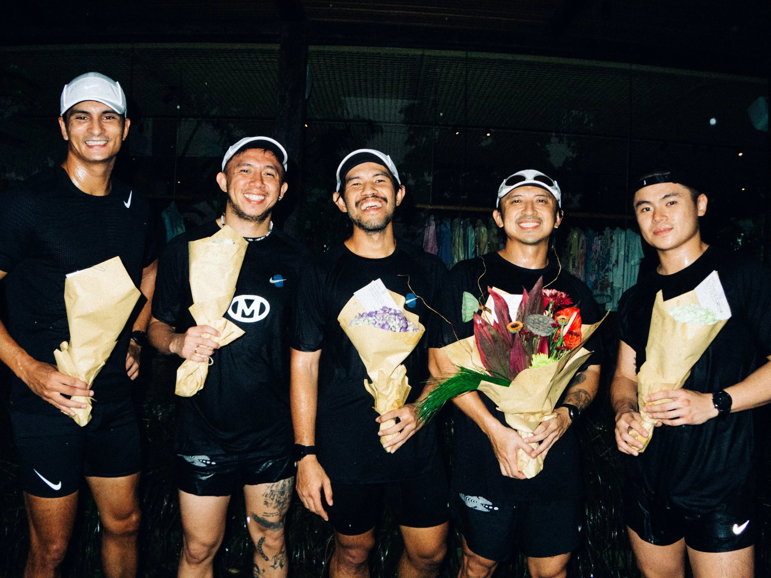 Five men in athletic clothing and caps holding bouquets of flowers, standing outdoors at night.