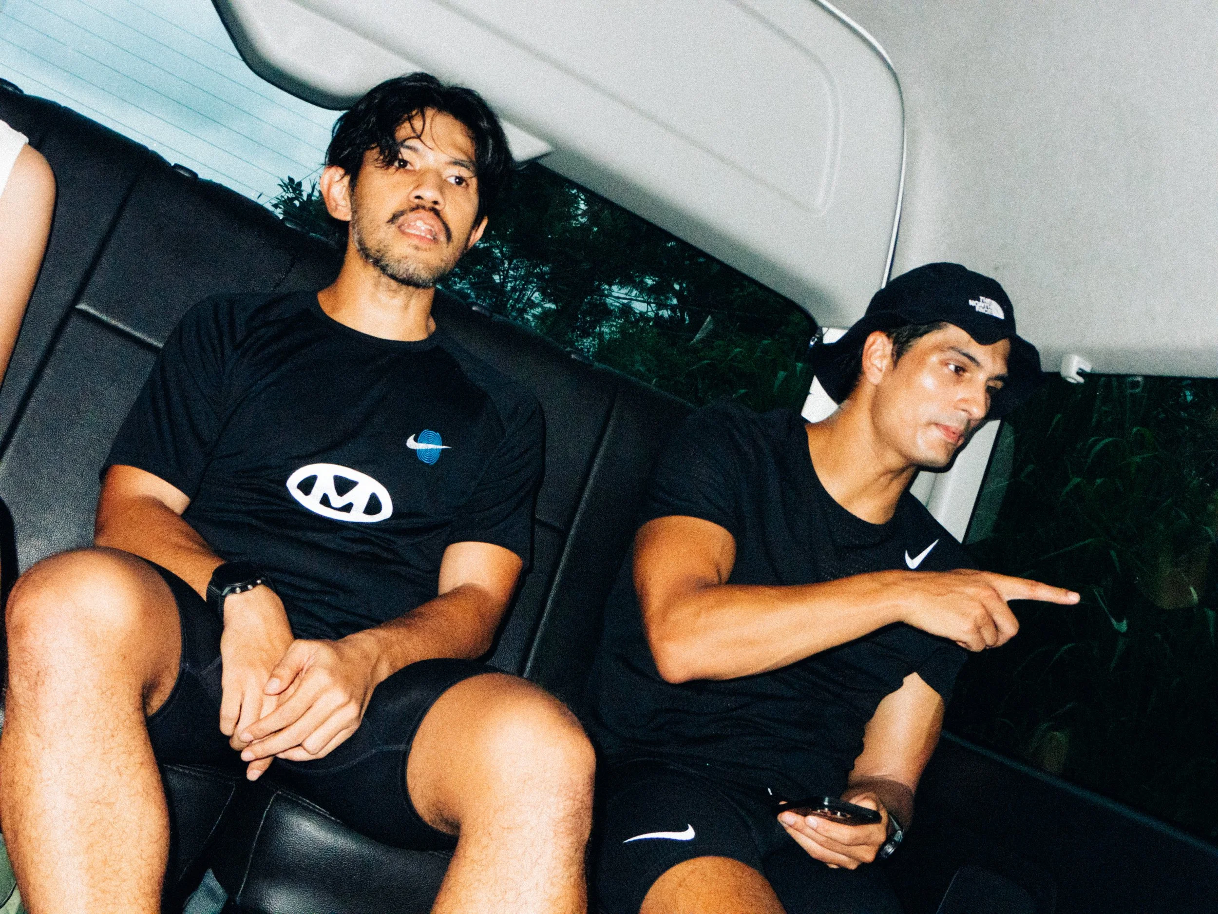 Two young men sitting on a bus in casual sportswear, one looking at the camera and the other looking at a phone