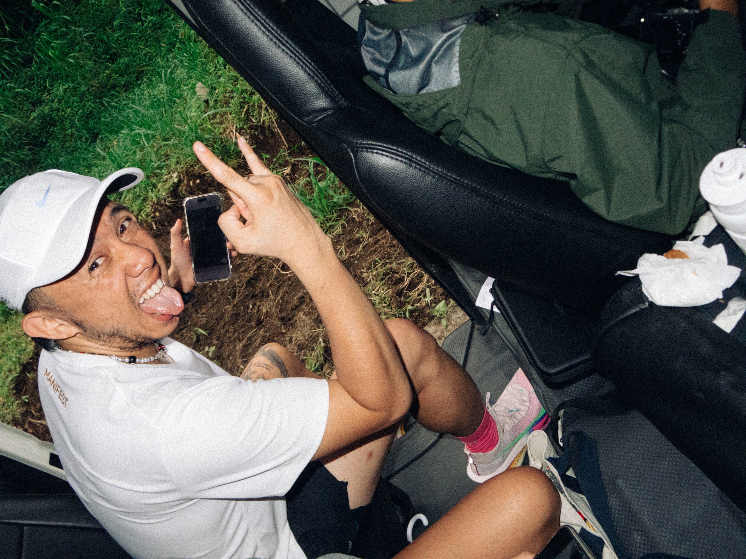 A person sitting in a golf cart, sticking out their tongue, wearing a white cap and t-shirt, holding a phone, making a peace sign with fingers, and surrounded by bags and miscellaneous items.