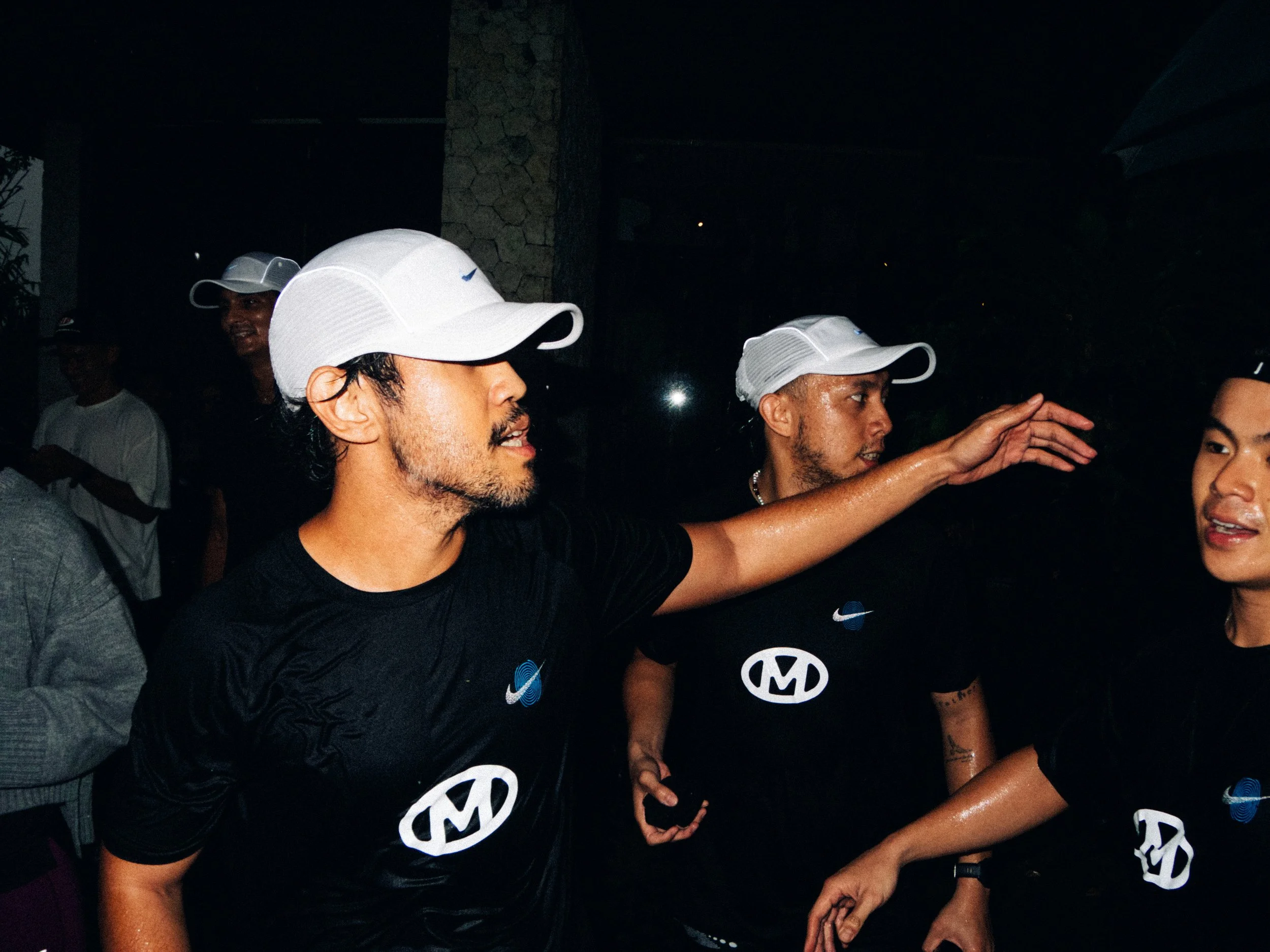 Three men wearing white caps and black athletic shirts, some with logos, engaging in a conversation or greeting at an outdoor event during nighttime.