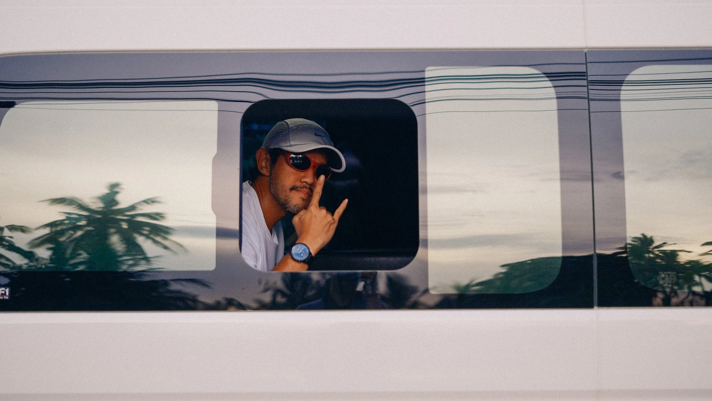 A man wearing sunglasses, a cap, and a watch, making a peace sign in a window of a vehicle, with palm trees and a cloudy sky reflected in the window.