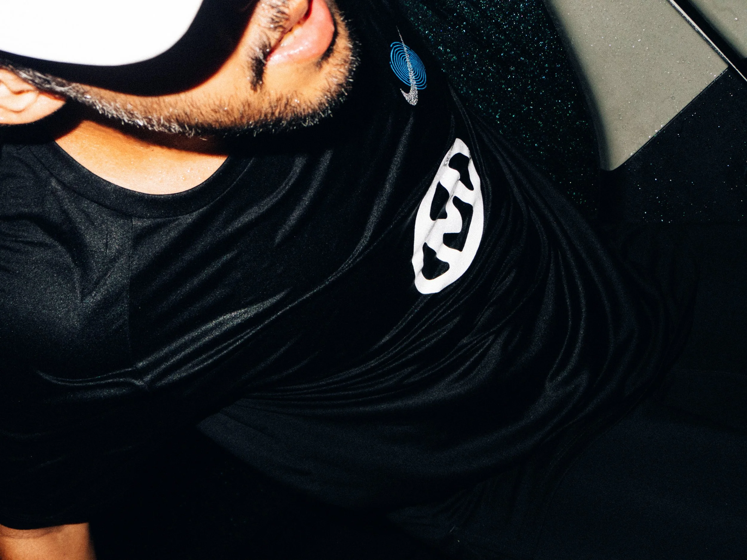 Close-up of a person wearing a black sports shirt with a white logo, sunglasses, and a beard, sitting on a dark bench.