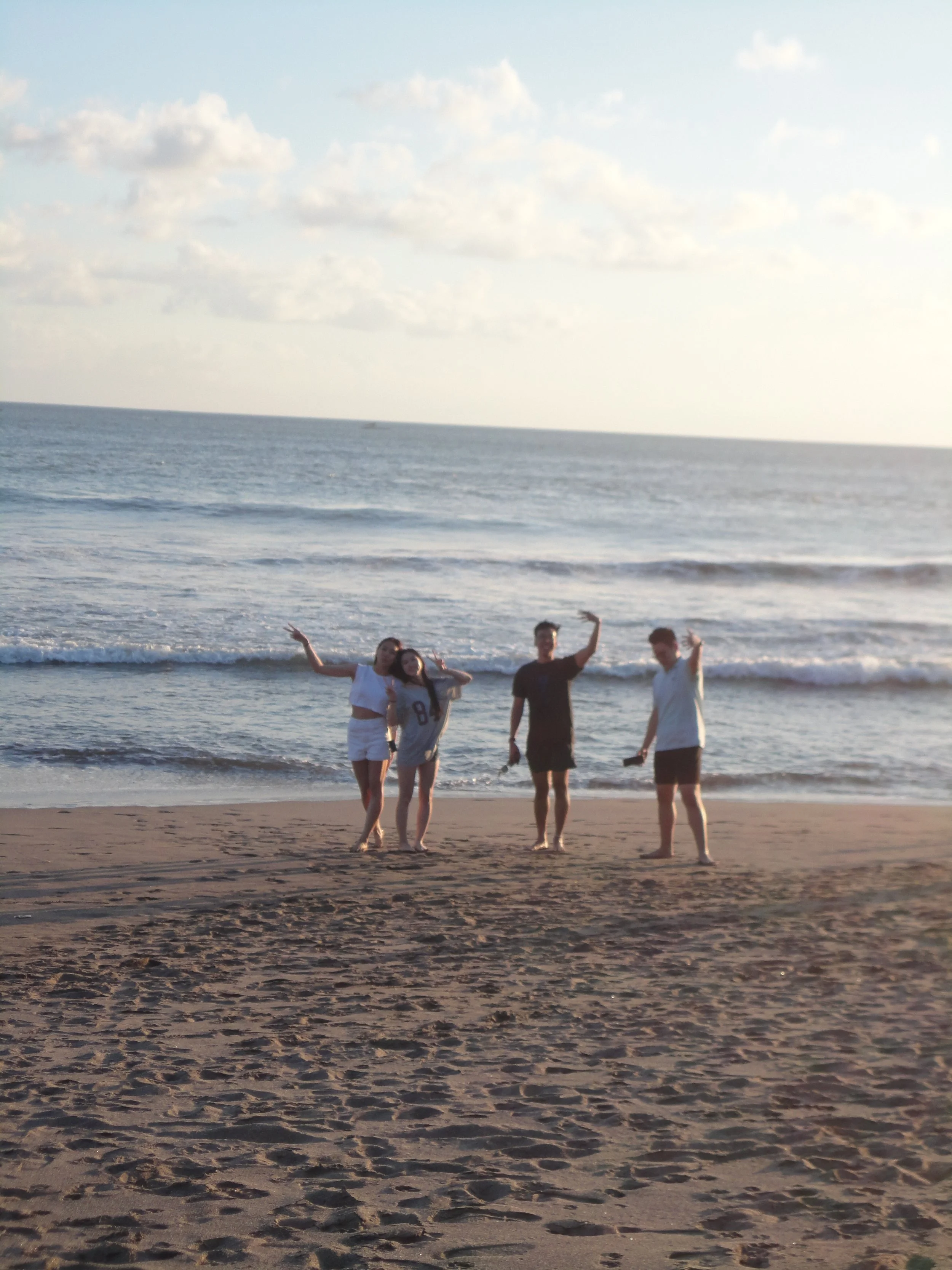Four people standing on the beach near the ocean, posing for a photo, with waves and a partly cloudy sky in the background.