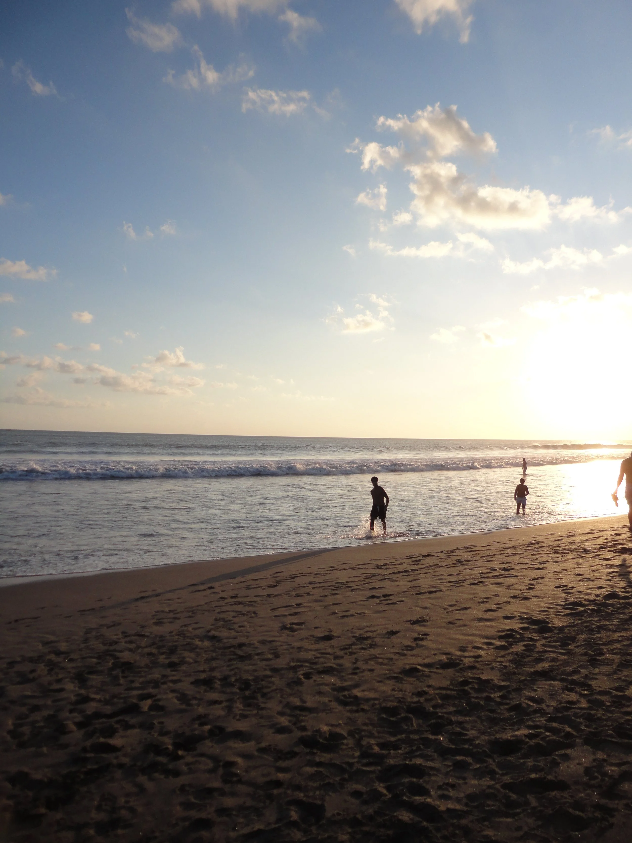 People enjoying the beach in the evening with the sun setting over the ocean, footprints in the sand, and clouds in the sky.
