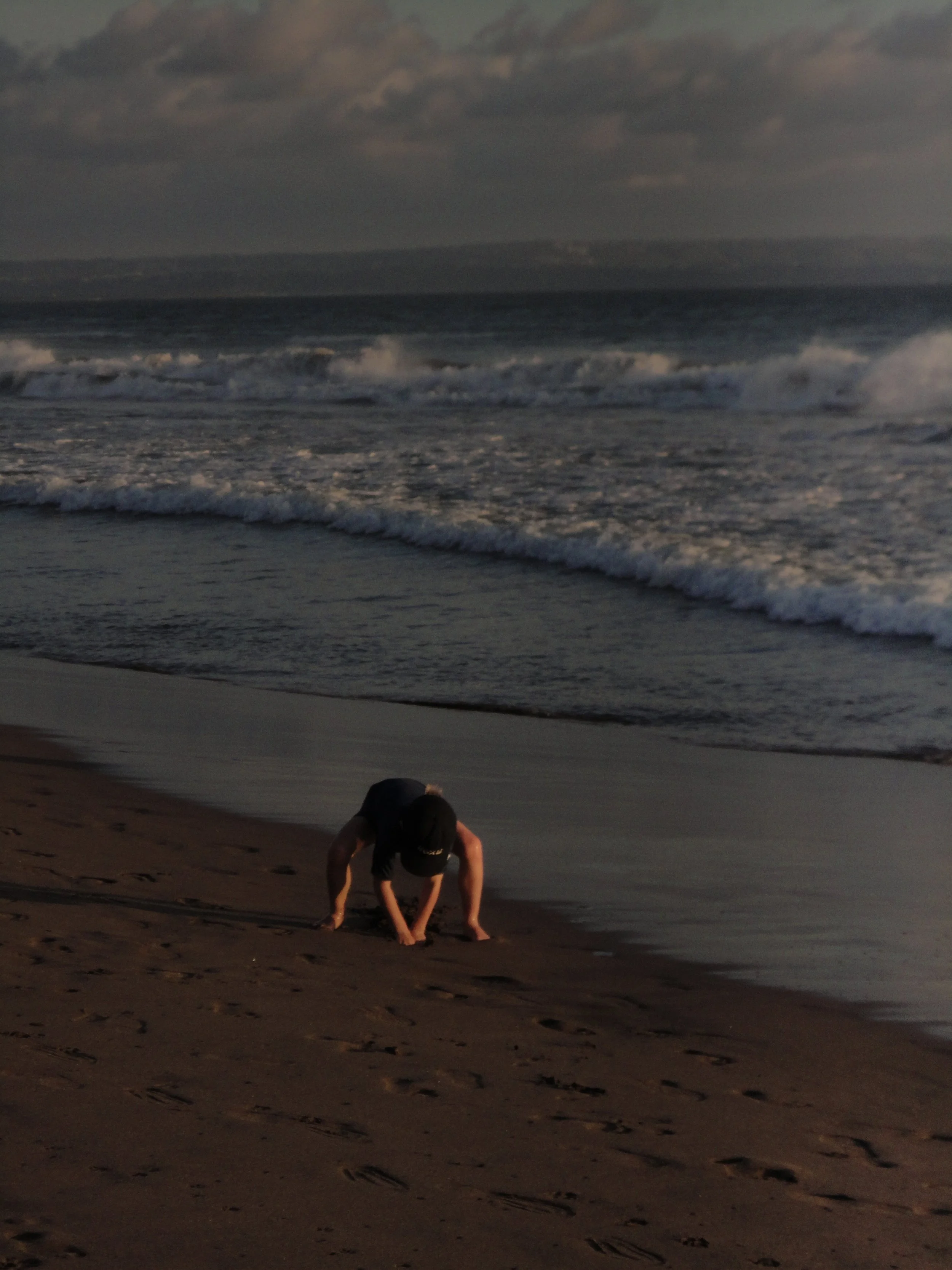 A person crouched on a sandy beach near the water's edge, with ocean waves and cloudy sky in the background.