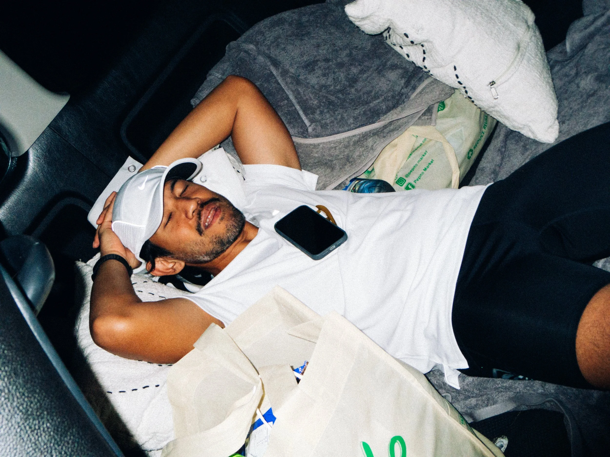 A man lying on a car floor sleeping or resting, wearing a white cap, white t-shirt, and black shorts, with a smartphone on his chest and surrounded by shopping bags and packages.