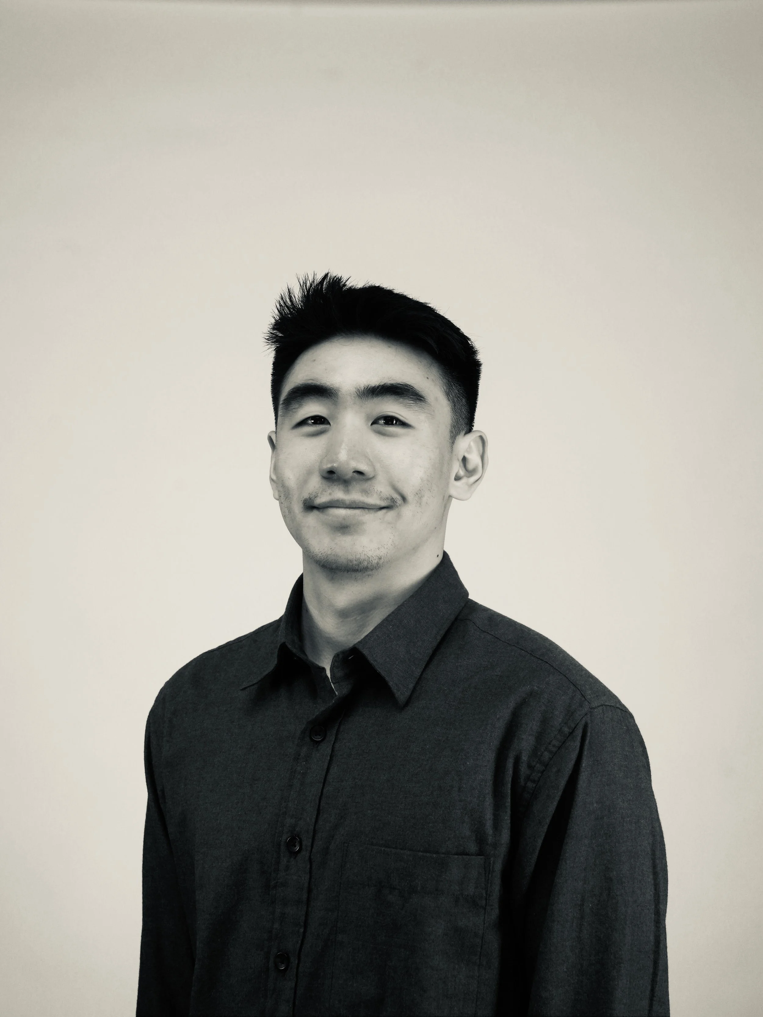 Black and white portrait of a young Asian man smiling, wearing a dark button-up shirt, standing against a plain light-colored background.