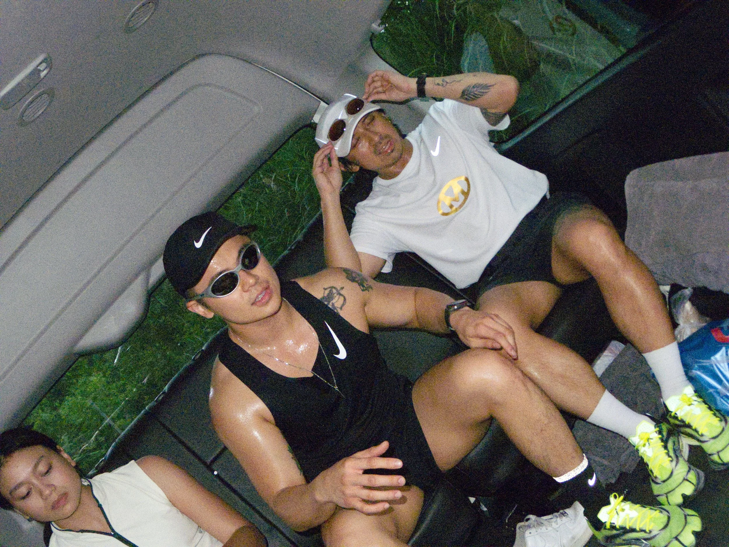 Two young men and a woman sitting on a bus, wearing athletic clothing and sunglasses, with wet skin and sweating.