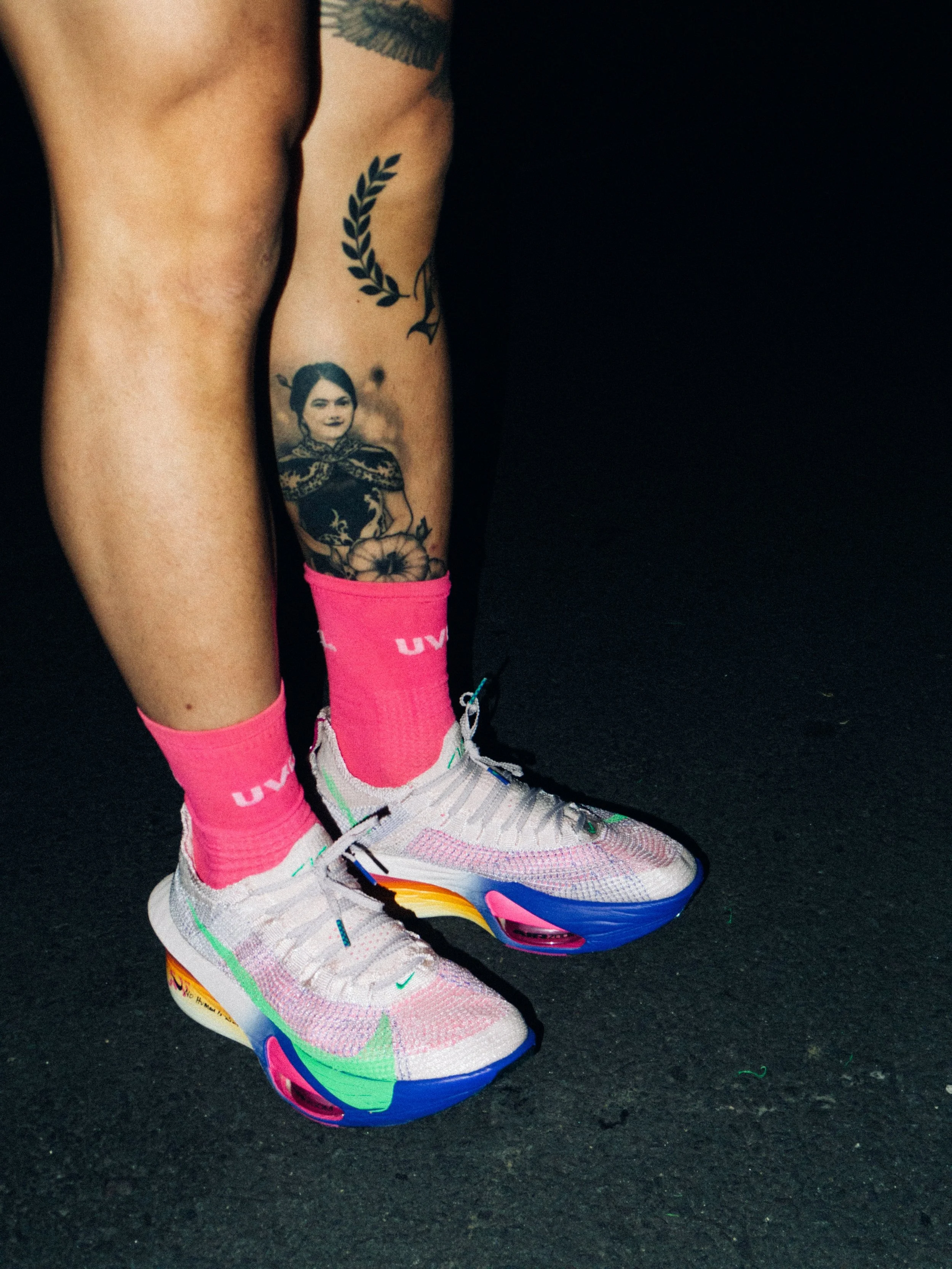 Close-up of person's legs, showing colorful running shoes, pink socks, and tattoos on the legs, against a black background.