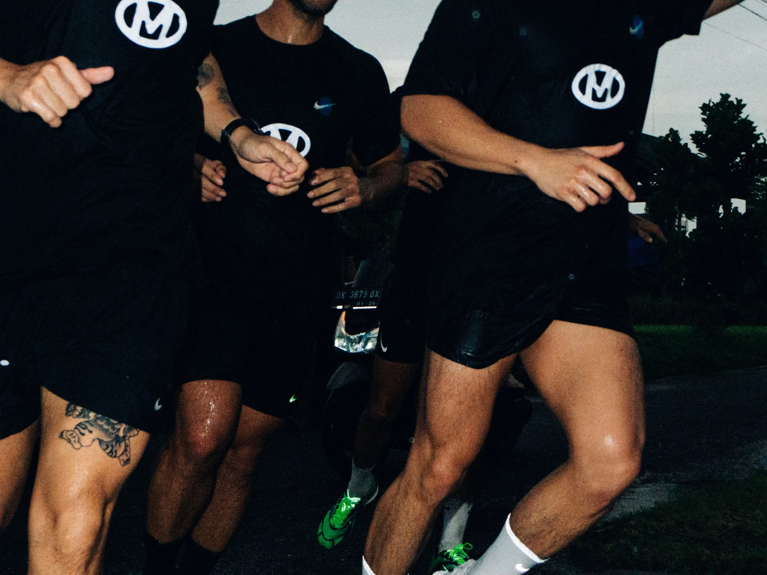 A group of runners wearing black athletic clothing, some with tattoos, running outdoors in rainy weather.