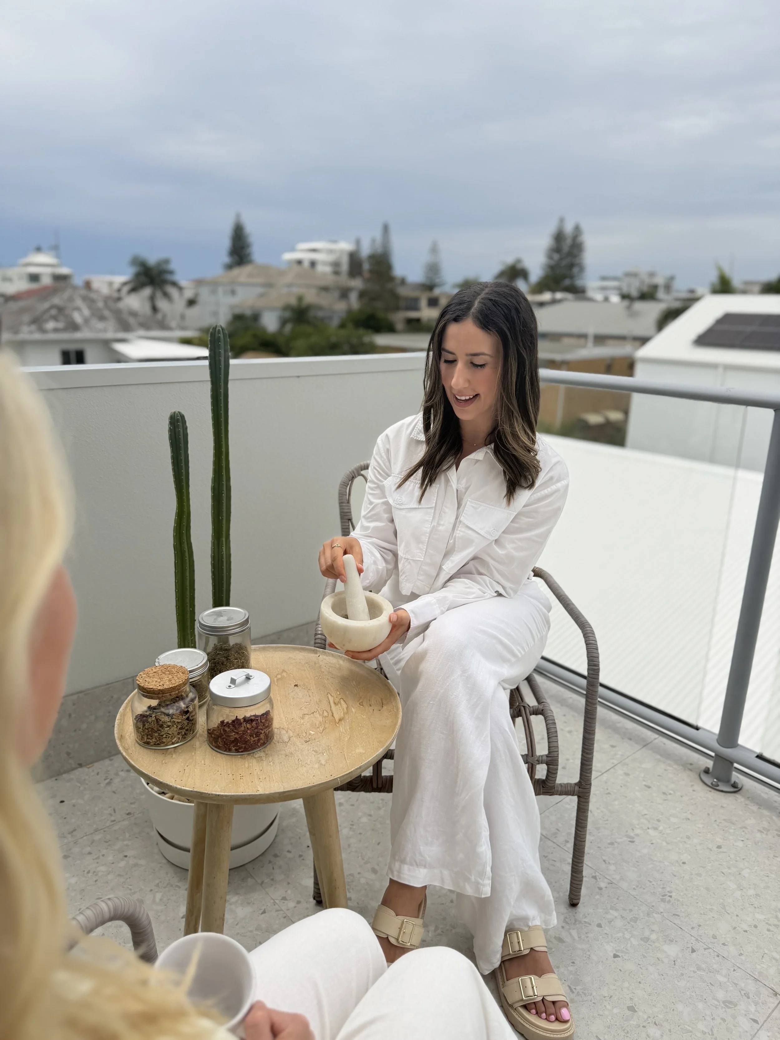 A woman in white attire sitting on a balcony, using a mortar and pestle, with jars of herbs on a small wooden table beside her. Behind her, there are tall cacti and a cityscape with residential buildings under a cloudy sky.