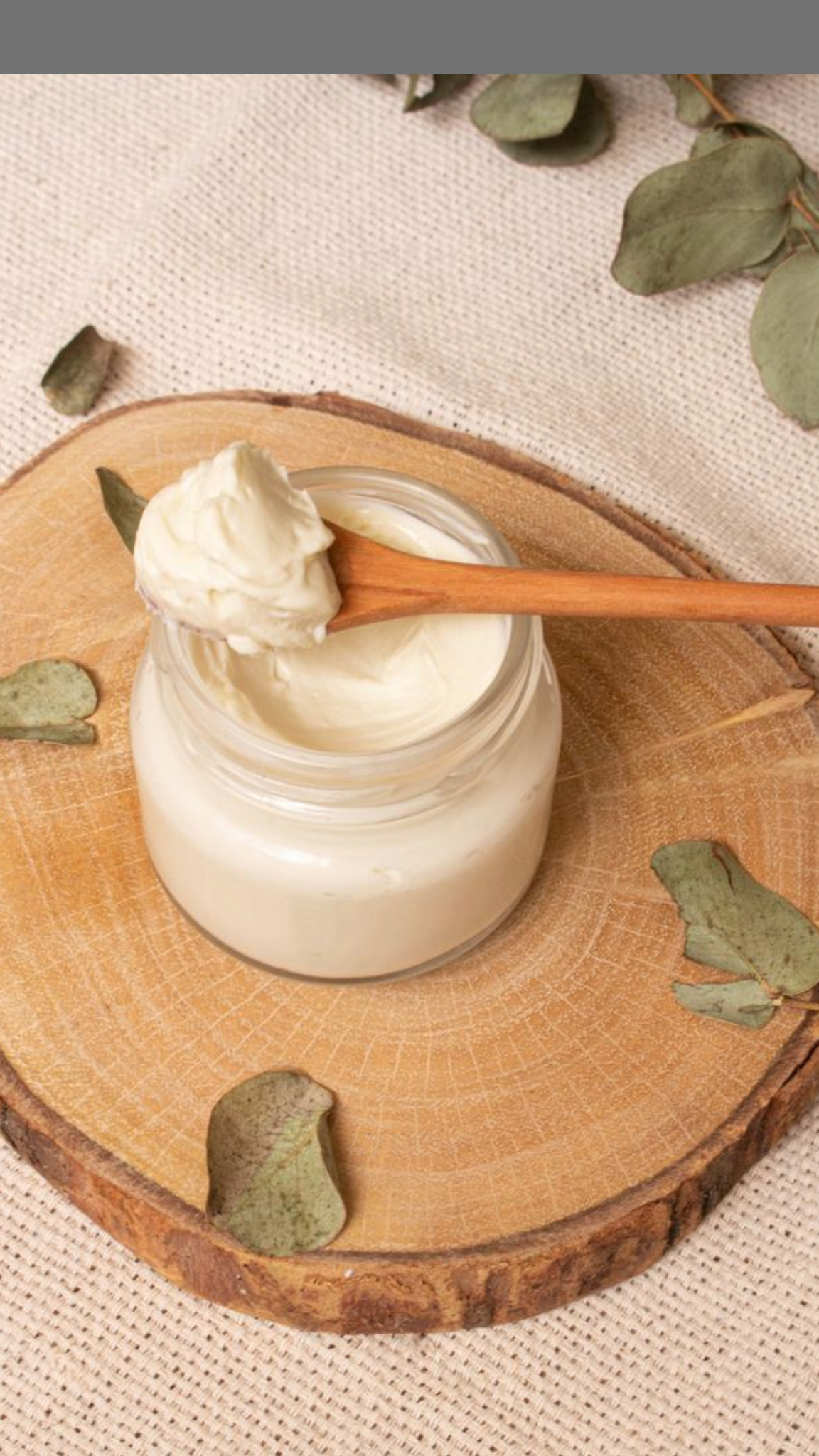 A glass jar filled with white cream or yogurt on a round wooden slab, with a wooden spoon resting on the jar and scooping out some of the cream. There are green leaves scattered around the jar on the wooden surface and a beige cloth in the background.