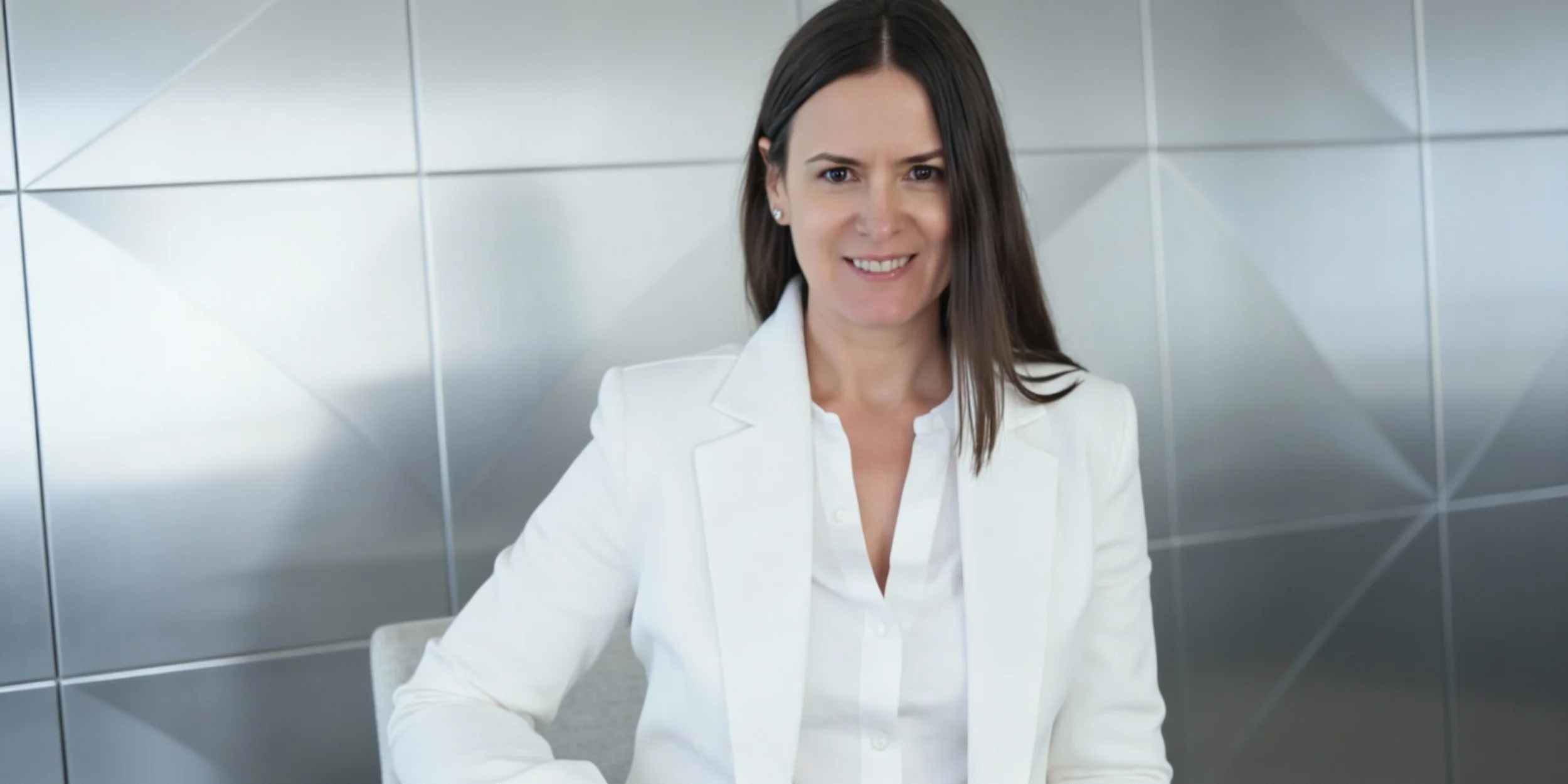 A woman with shoulder-length dark hair wearing a white blazer and white blouse, sitting in front of a metallic, geometric-patterned wall, smiling at the camera.