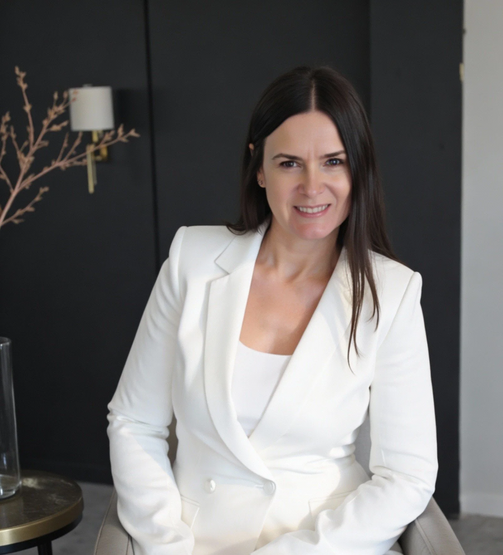 A woman with long dark hair wearing a white blazer and matching top, sitting on a chair in a modern office setting with black walls and a decorative lamp in the background.