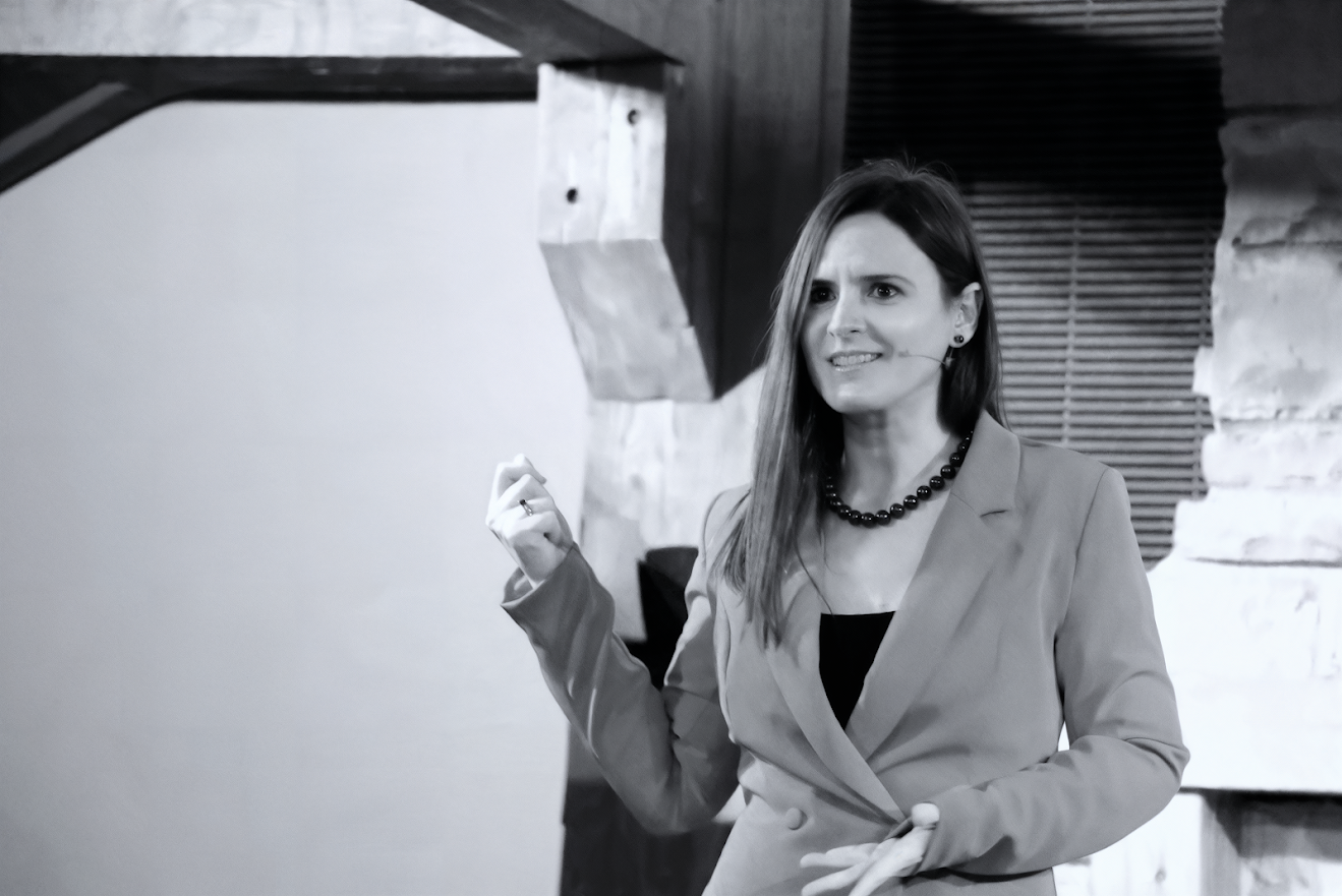 A woman speaking or presenting, wearing a blazer and a necklace, with an expressive facial expression, standing indoors against a background with wooden and brick elements.