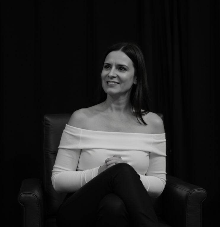 A smiling woman with shoulder-length dark hair, sitting on a chair with her hands clasped, wearing an off-the-shoulder white top and dark pants, in front of dark curtains.