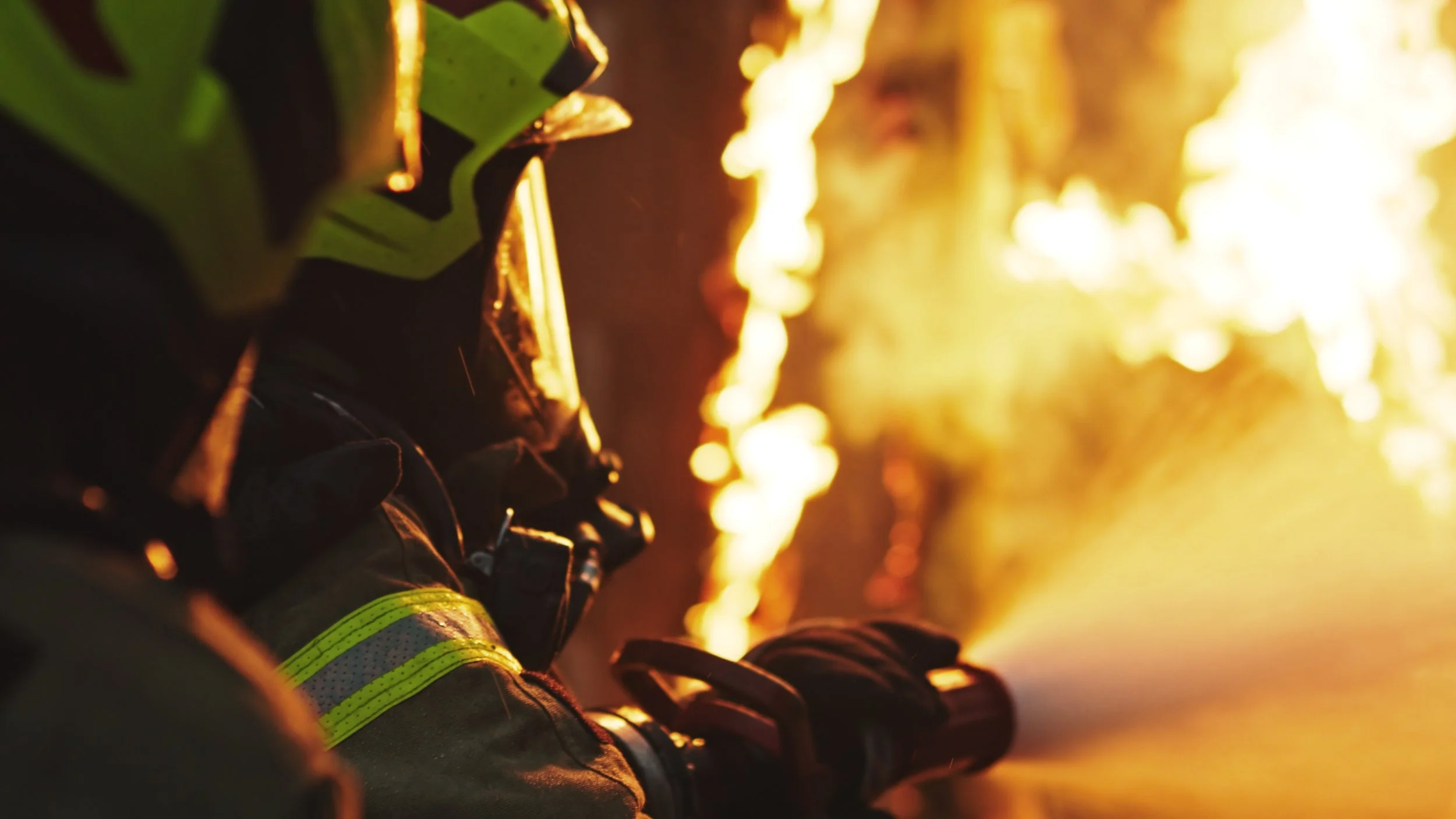 Firefighter in protective gear fighting a large fire with flames in the background.