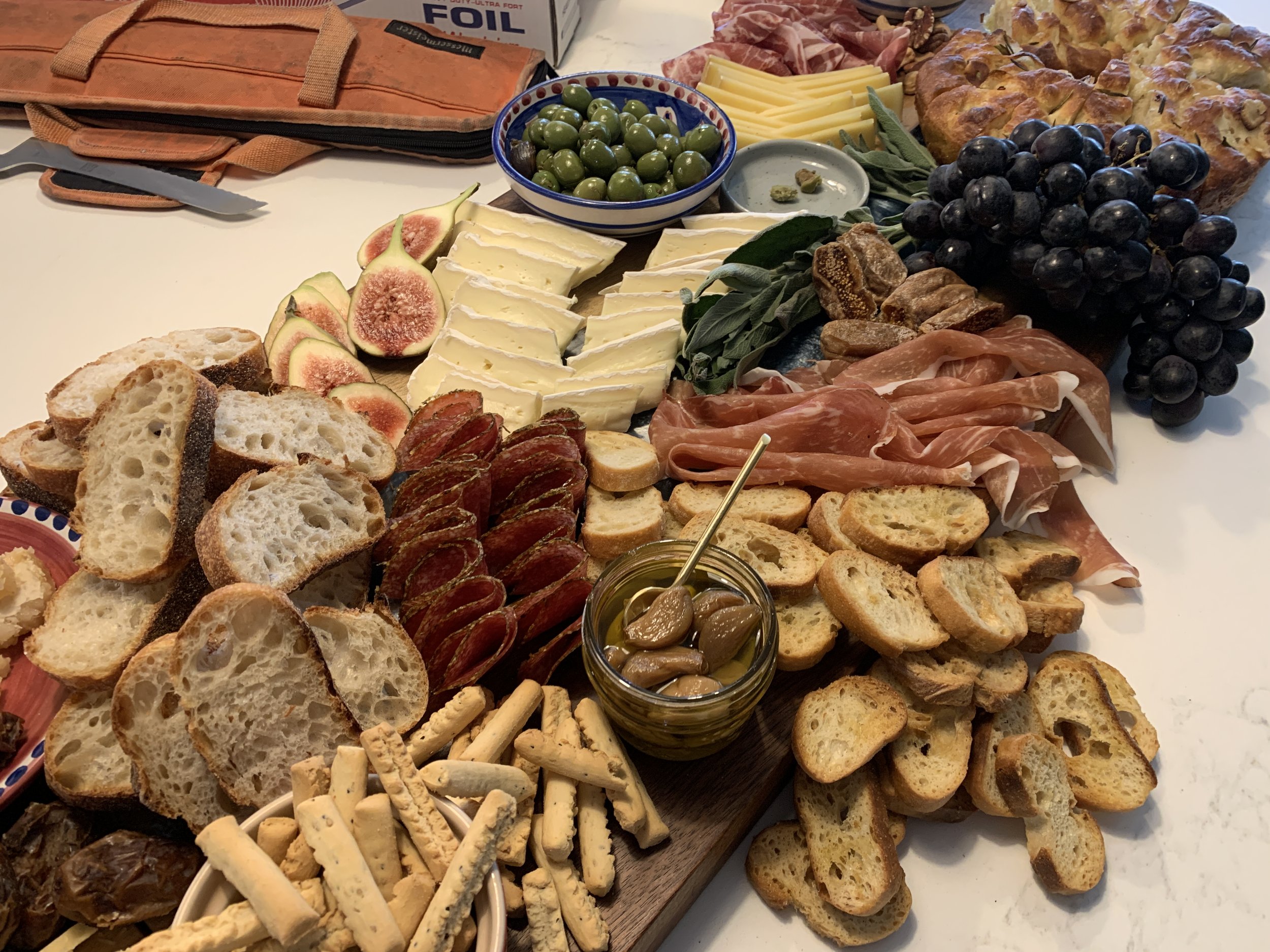 A spread of assorted cheeses, sliced bread, cured meats, grapes, figs, olives, and breadsticks on a table for a charcuterie board or appetizer platter.