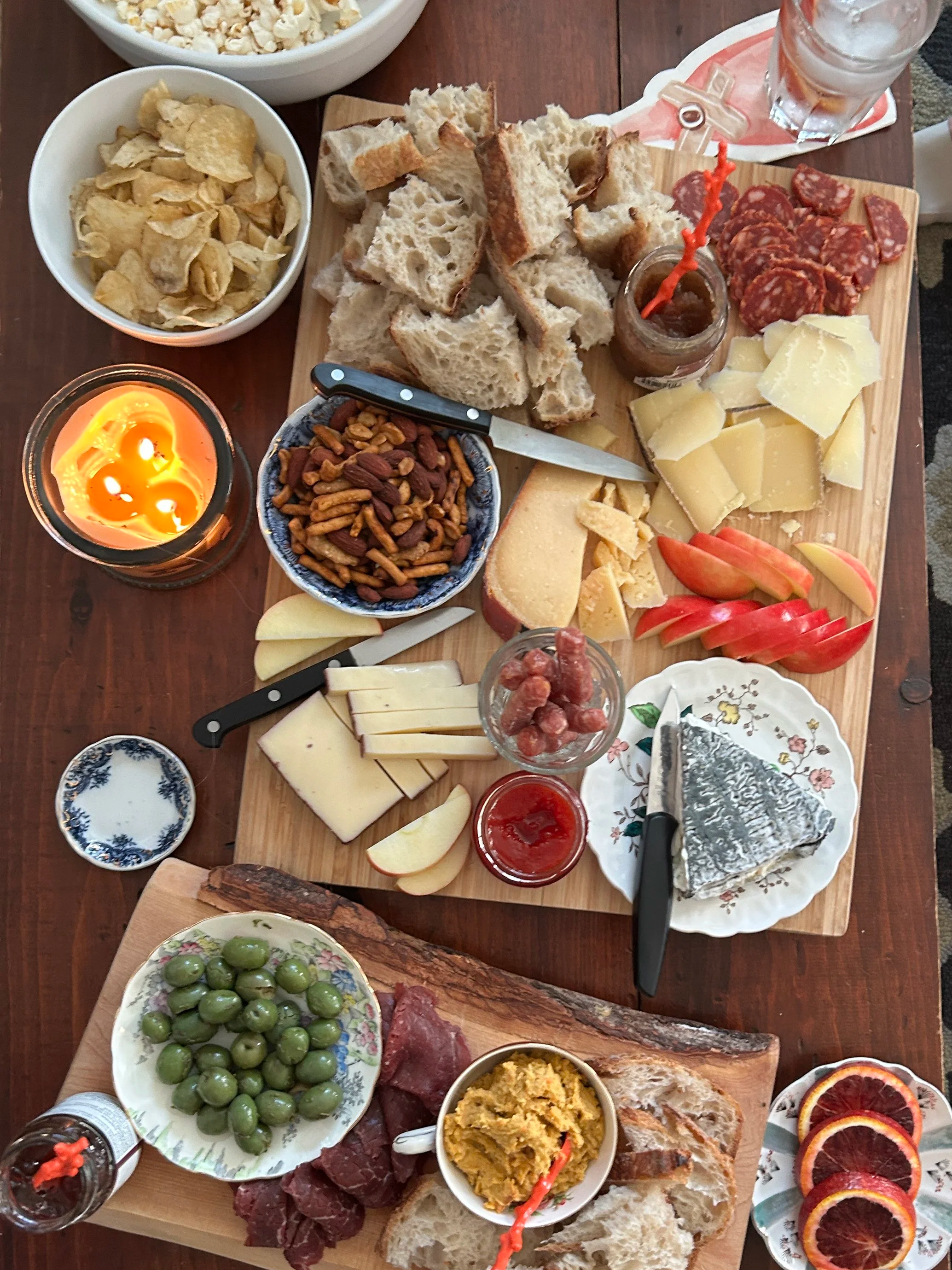 A charcuterie board with bread, cheeses, meats, olives, nuts, and fruits, set on a wooden table, with bowls of popcorn and potato chips and a candle nearby.