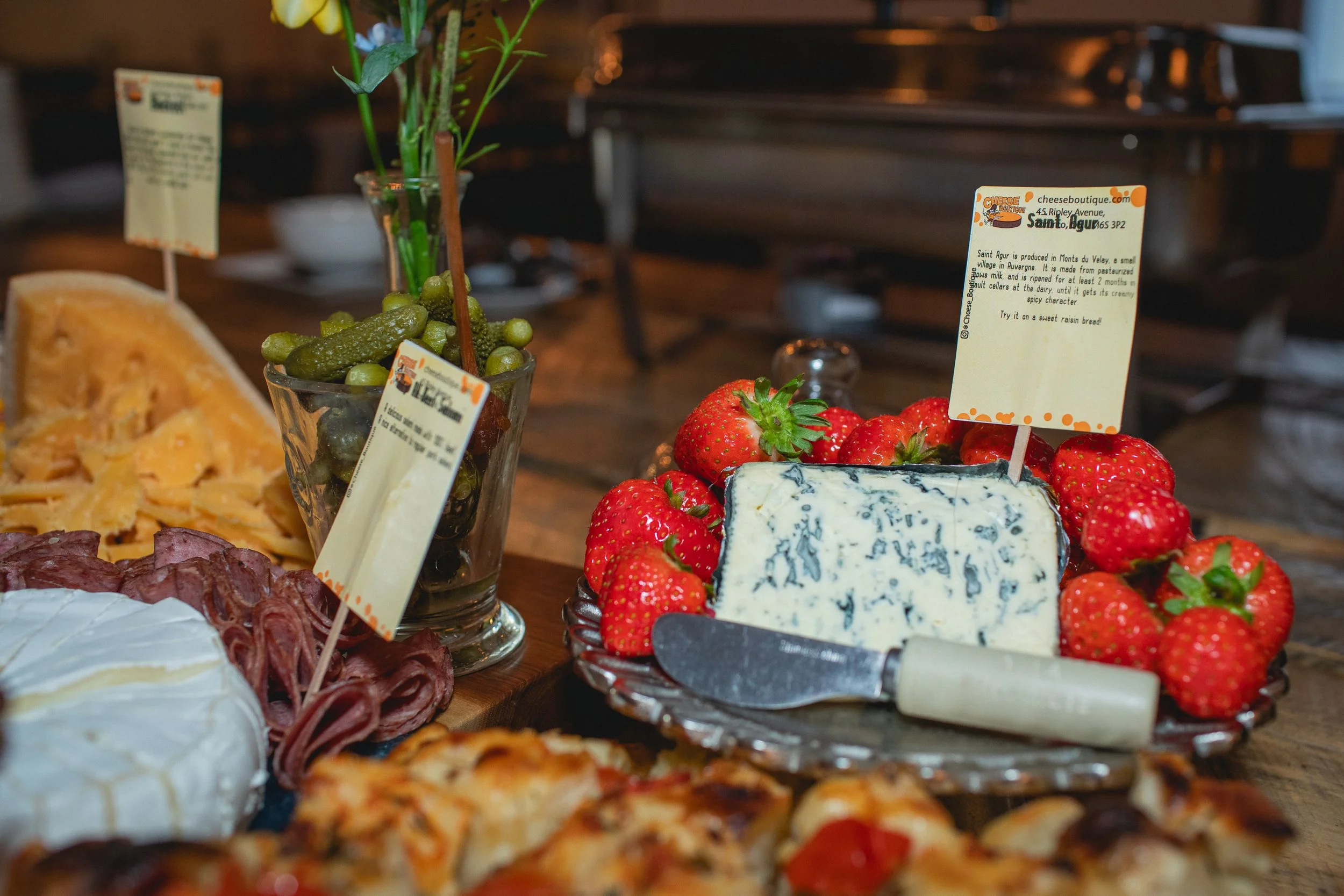 A cheese platter with strawberries, blue cheese, and a cheese knife, surrounded by various cheeses, slices of cured meat, and a bowl of pickles, all on a wooden table.