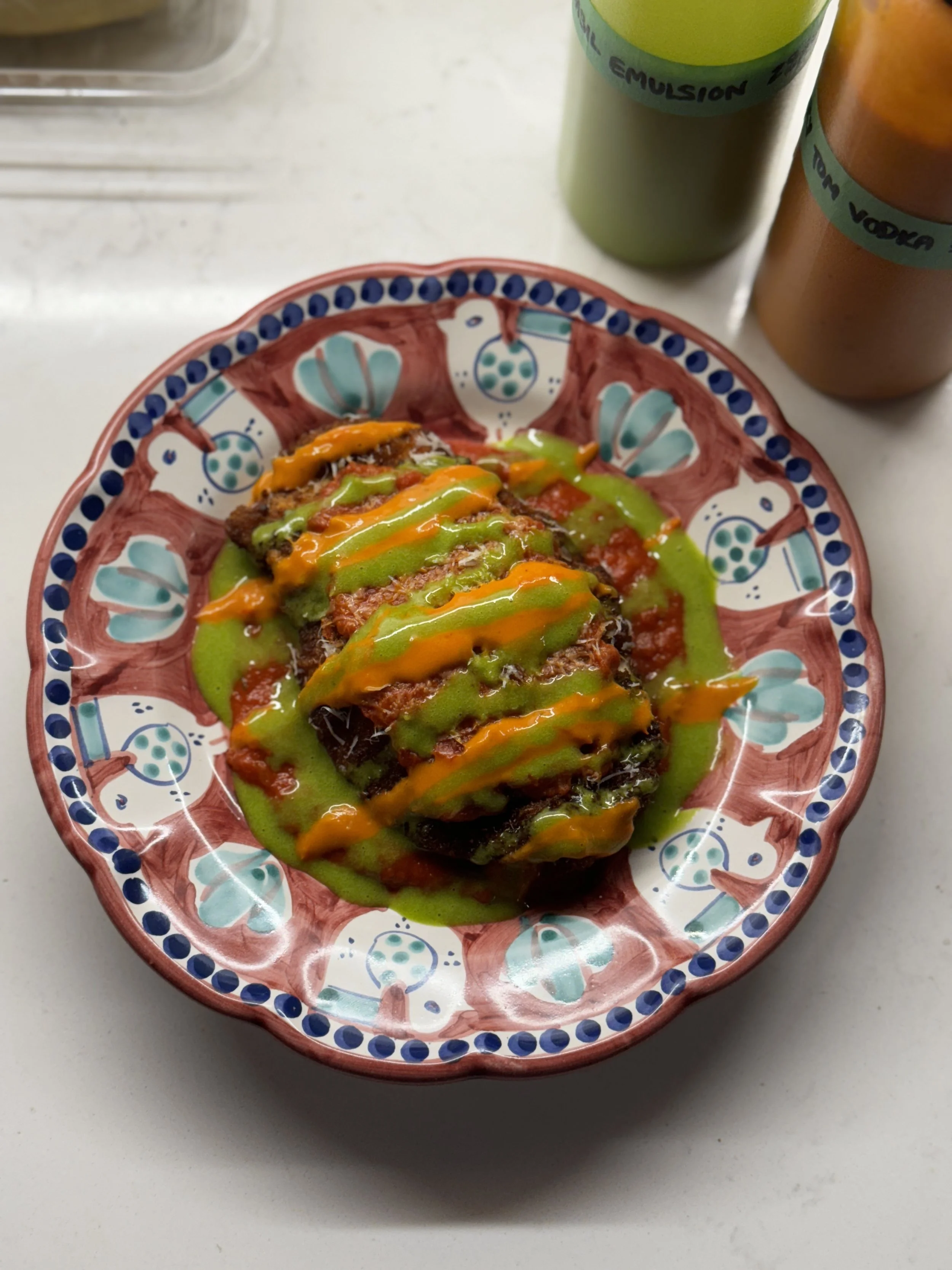 An Italian Vietri dish plated with a piece of chicken parmesan, drizzled with a basil emulsion and cherry tomato vodka sauce