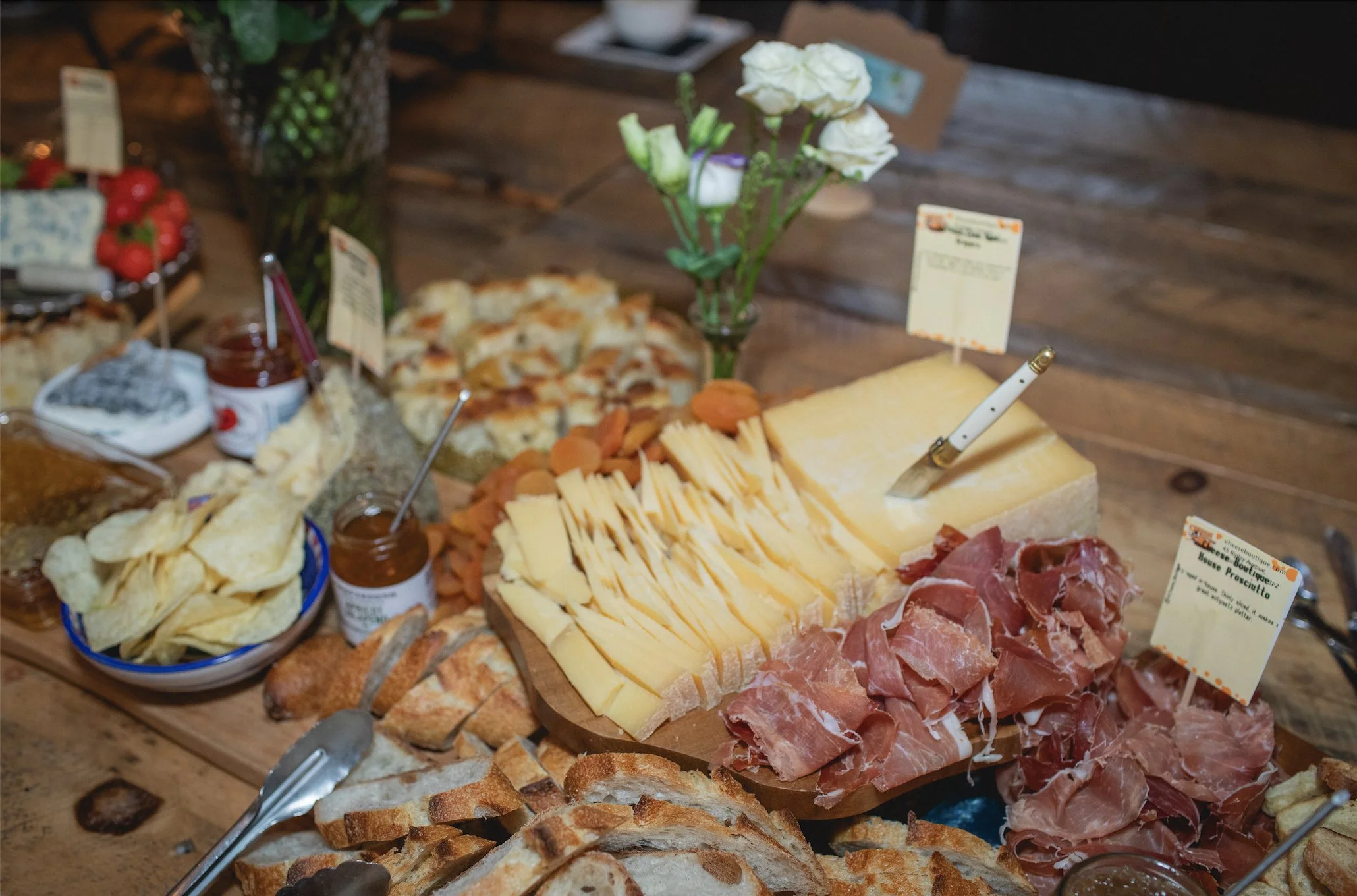 Cheese and prosciutto platter on a wooden table with sliced bread, potato chips, jams, and a vase of white flowers.