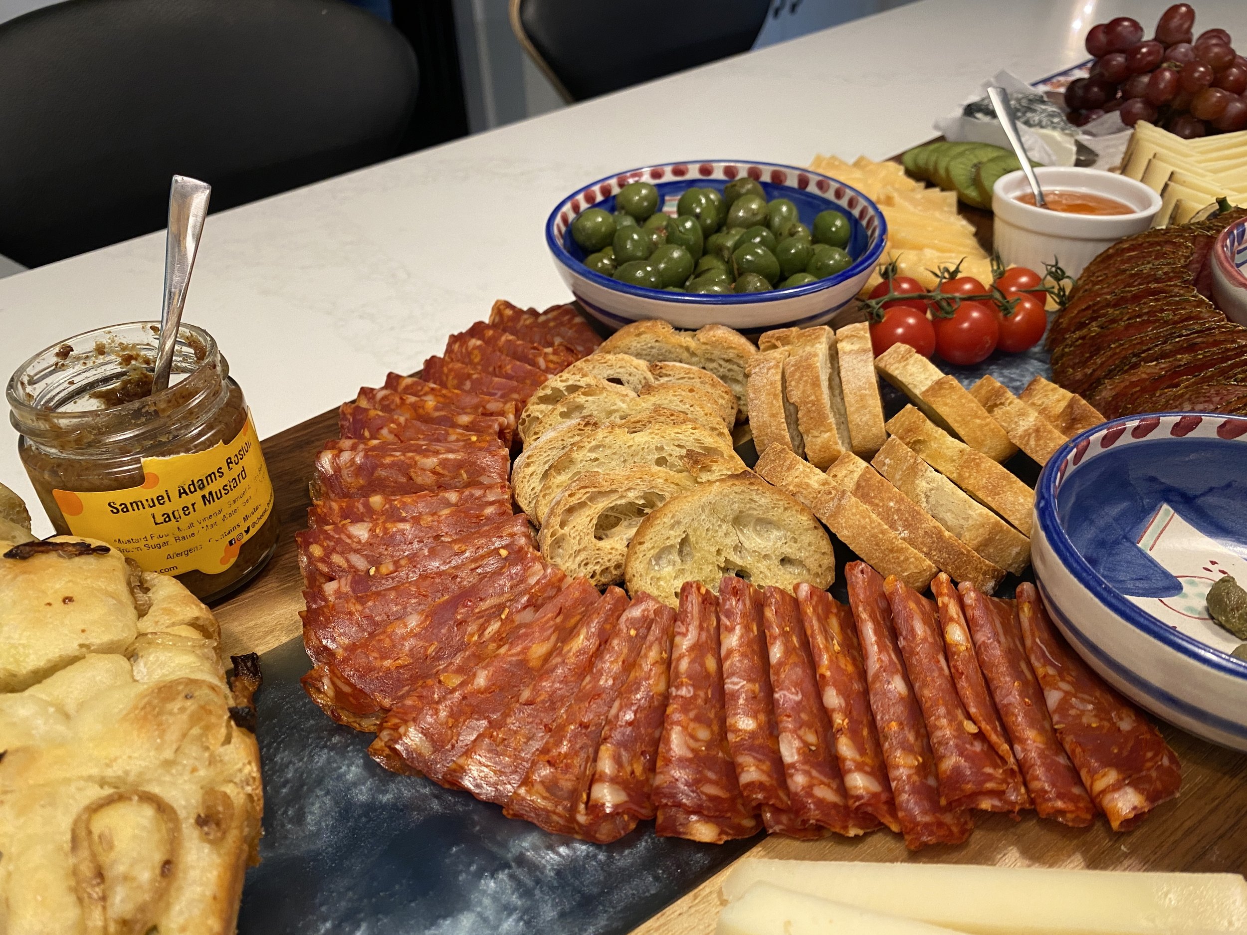A charcuterie board with slices of salami, baguette slices, green olives, cherry tomatoes, grapes, cheese, and small bowls of sauces on a white table.