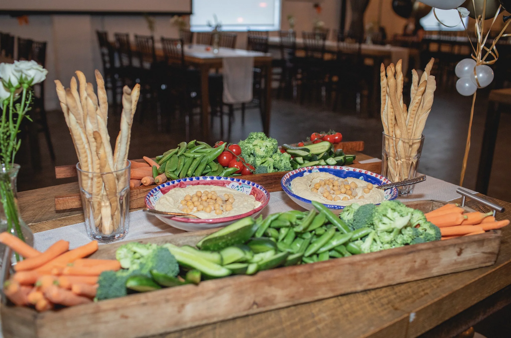 A wooden table with trays of fresh vegetables, bowls of hummus with chickpeas, and breadsticks in glasses at a party or gathering.