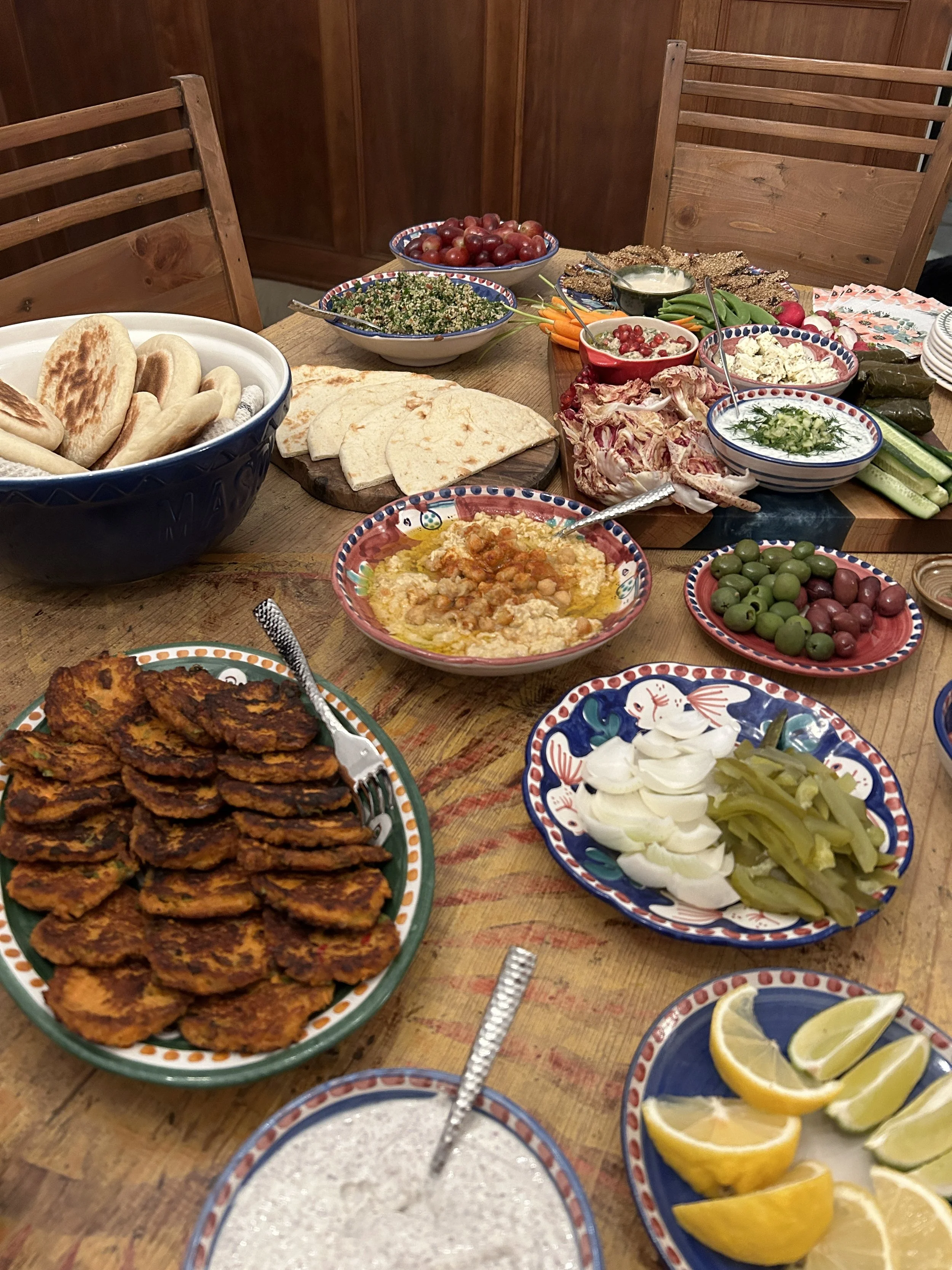 A table set with various traditional Middle Eastern dishes, including falafel, hummus, tabbouleh, pickles, olives, sliced lemons, cucumber slices, grape leaves, and a bowl of pomegranate seeds, with bread and other side dishes.