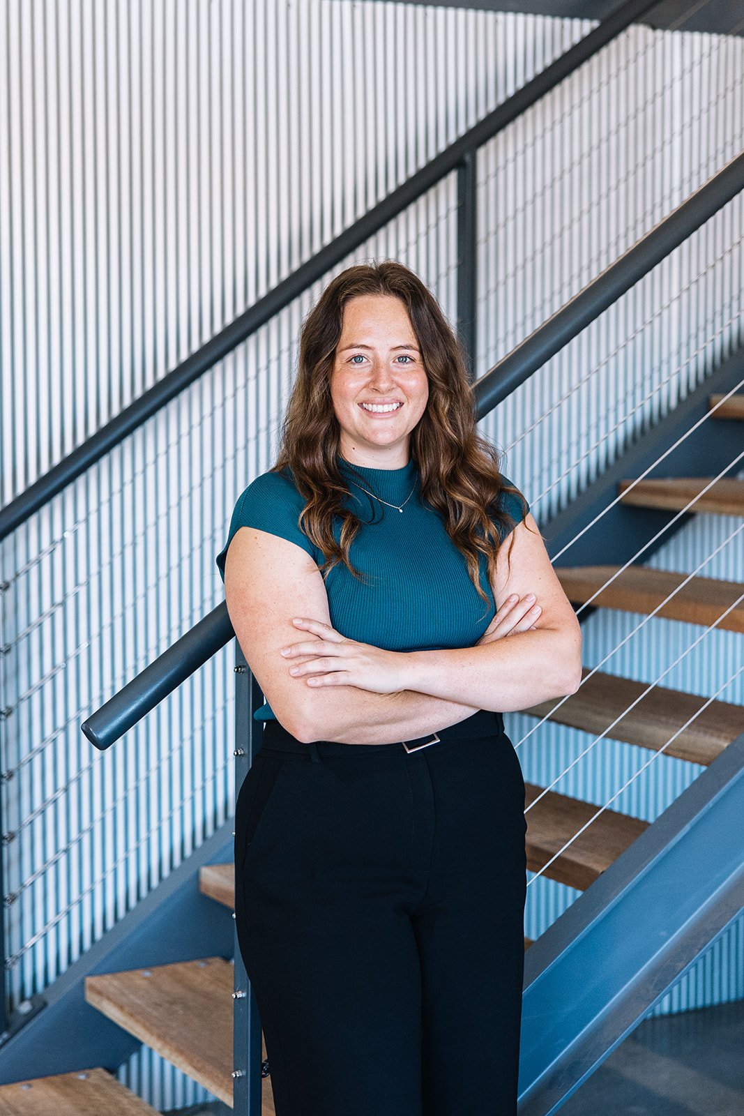 A woman with long, wavy brown hair in a teal short-sleeve top and black pants standing on a staircase with metal railings and wooden steps, smiling with arms crossed.