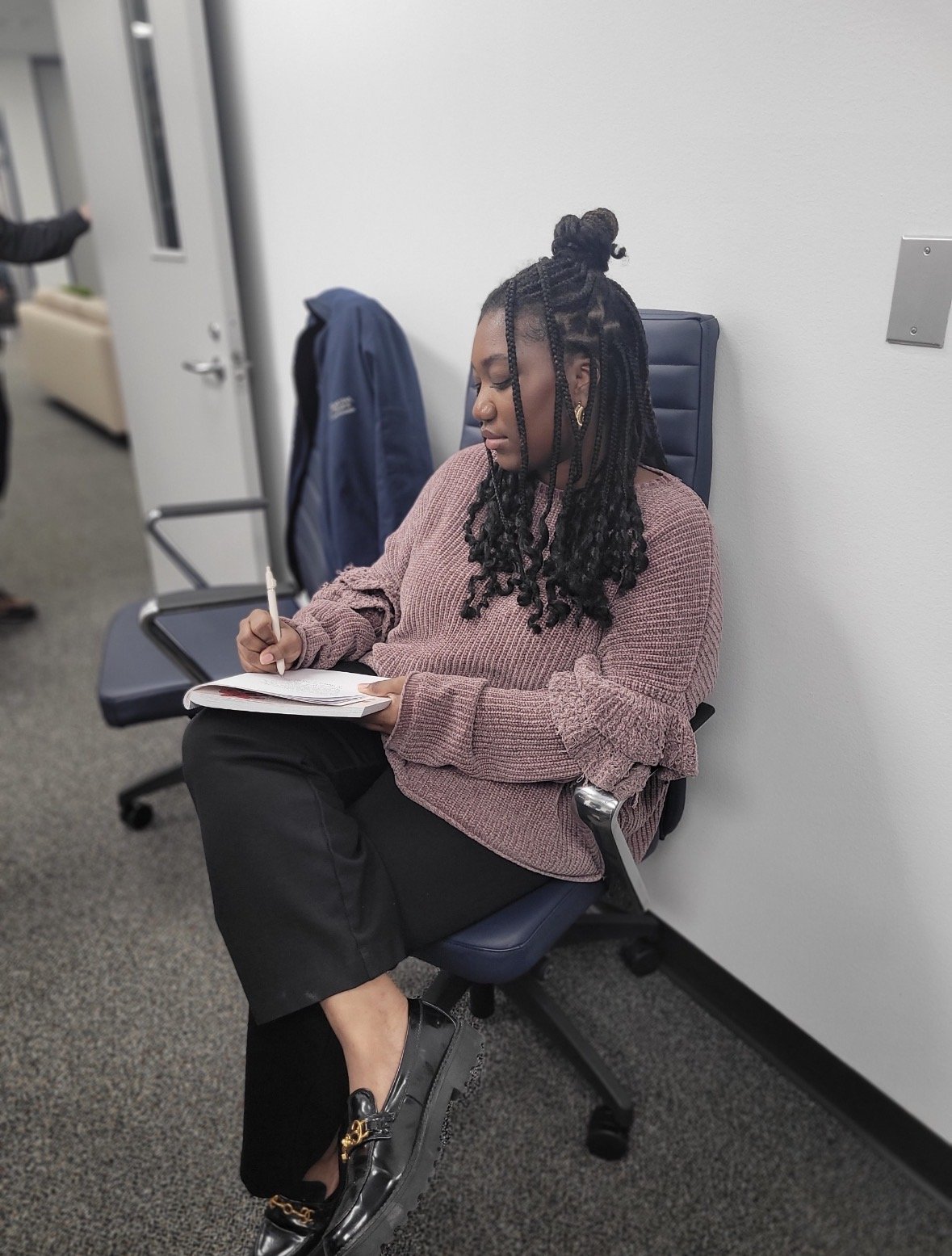 A woman sitting on an office chair, writing in a notebook with a pen, wearing a pink sweater, black pants, and shiny black loafers.