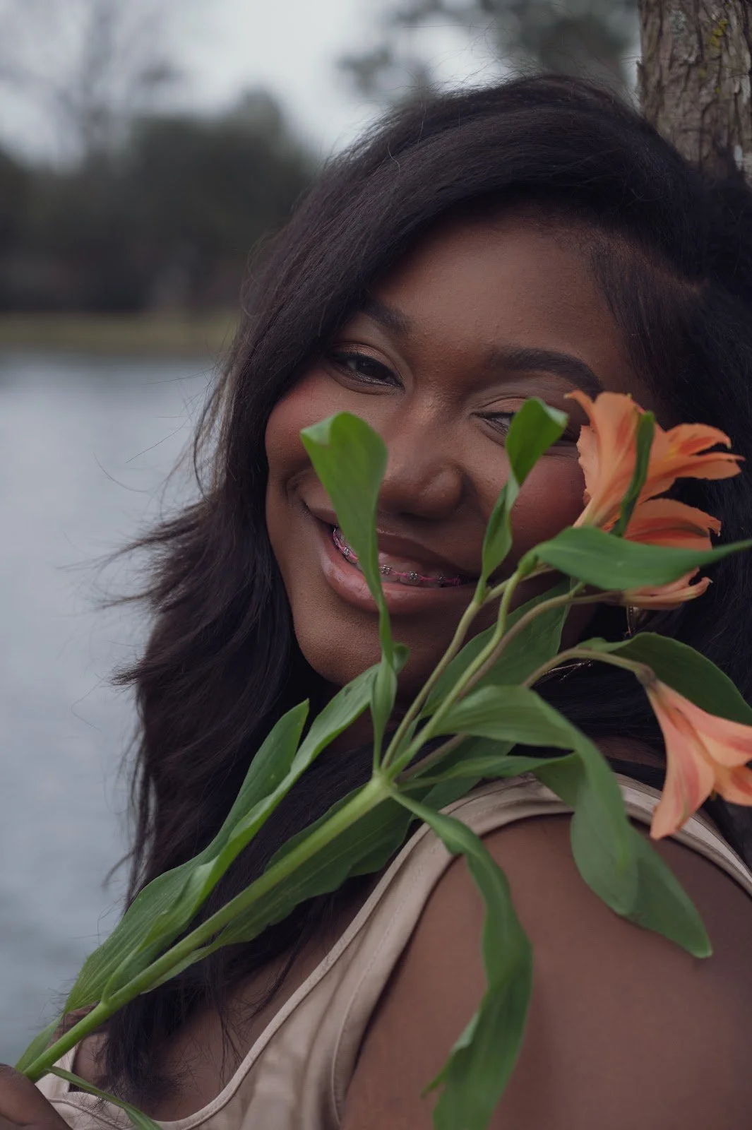A woman with dark hair and braces smiling while holding peach-colored flowers near her face.