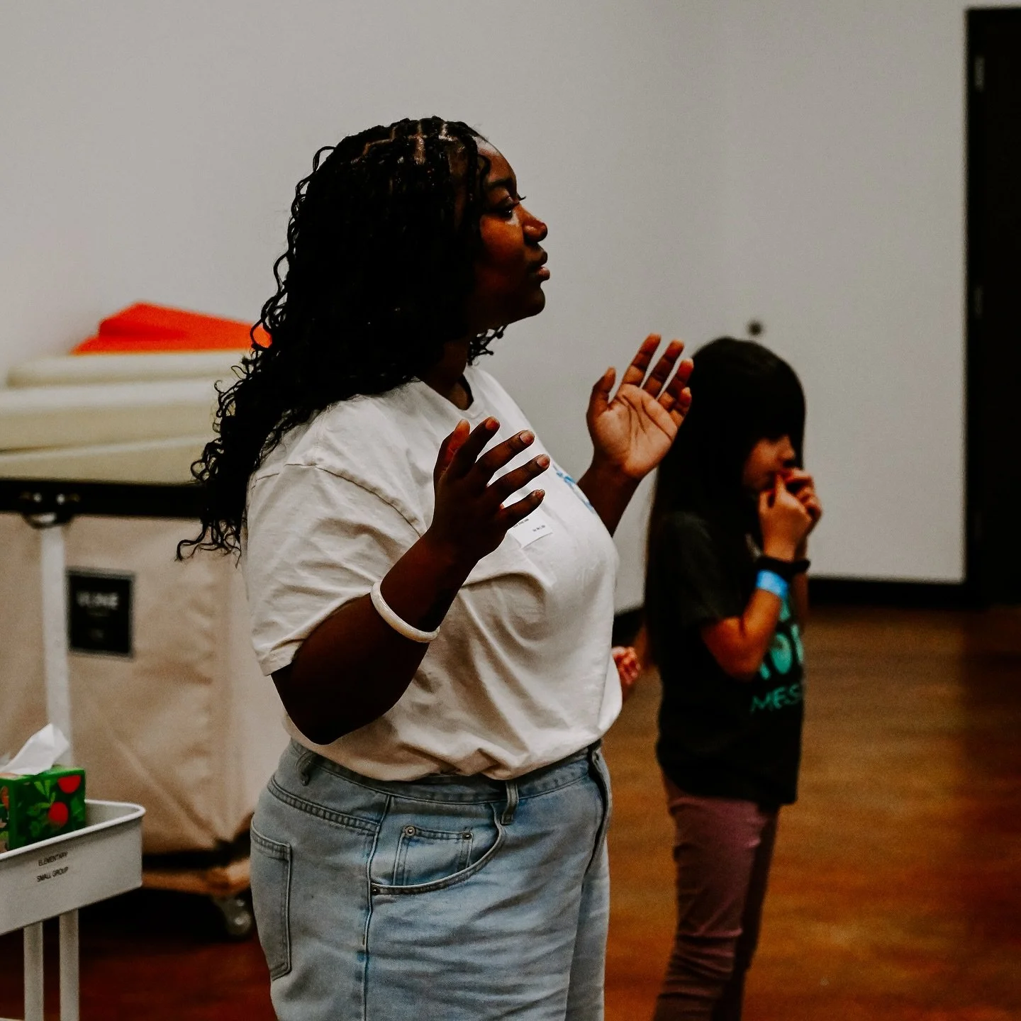 A woman with curly hair and wearing a white t-shirt is standing with her hands raised, seemingly speaking or explaining something. A young girl is standing nearby, holding her nose or mouth, in a room with wooden floors and a plain white wall.