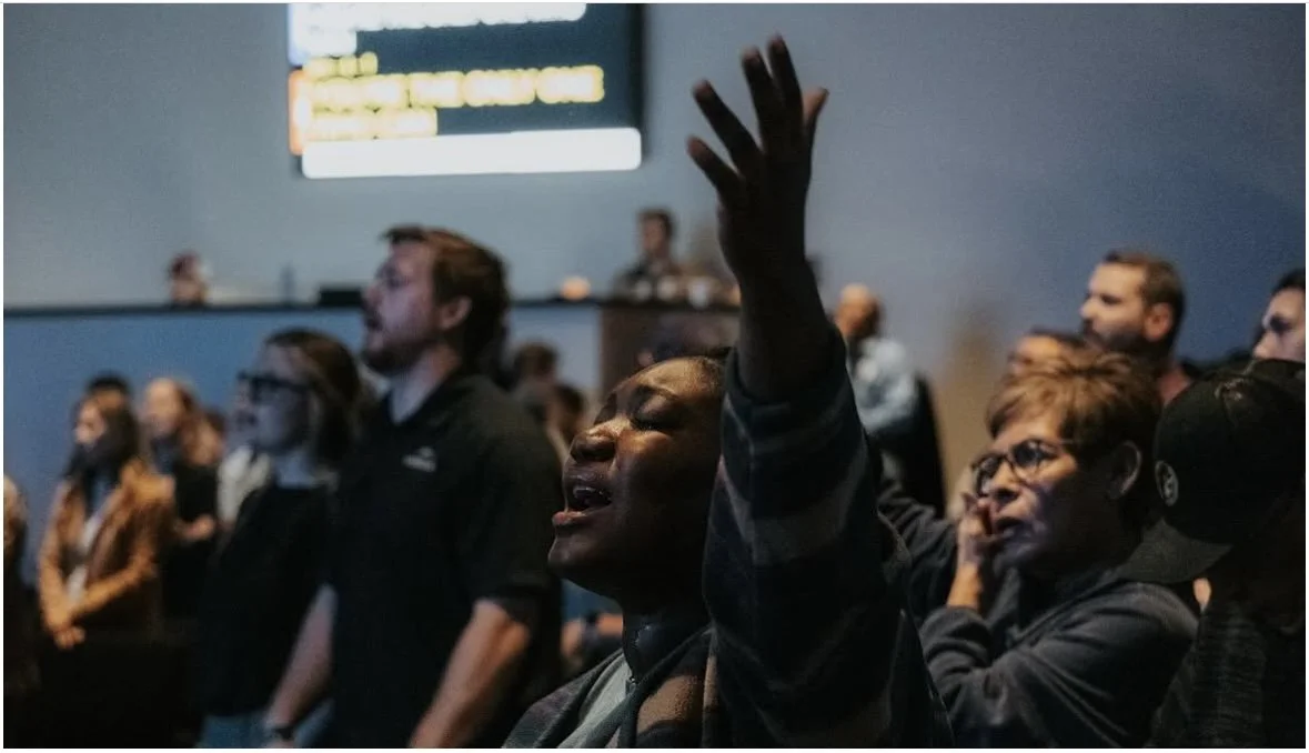 A group of people in a religious or worship setting, with a woman in the foreground raising her hand and eyes closed in prayer or worship.