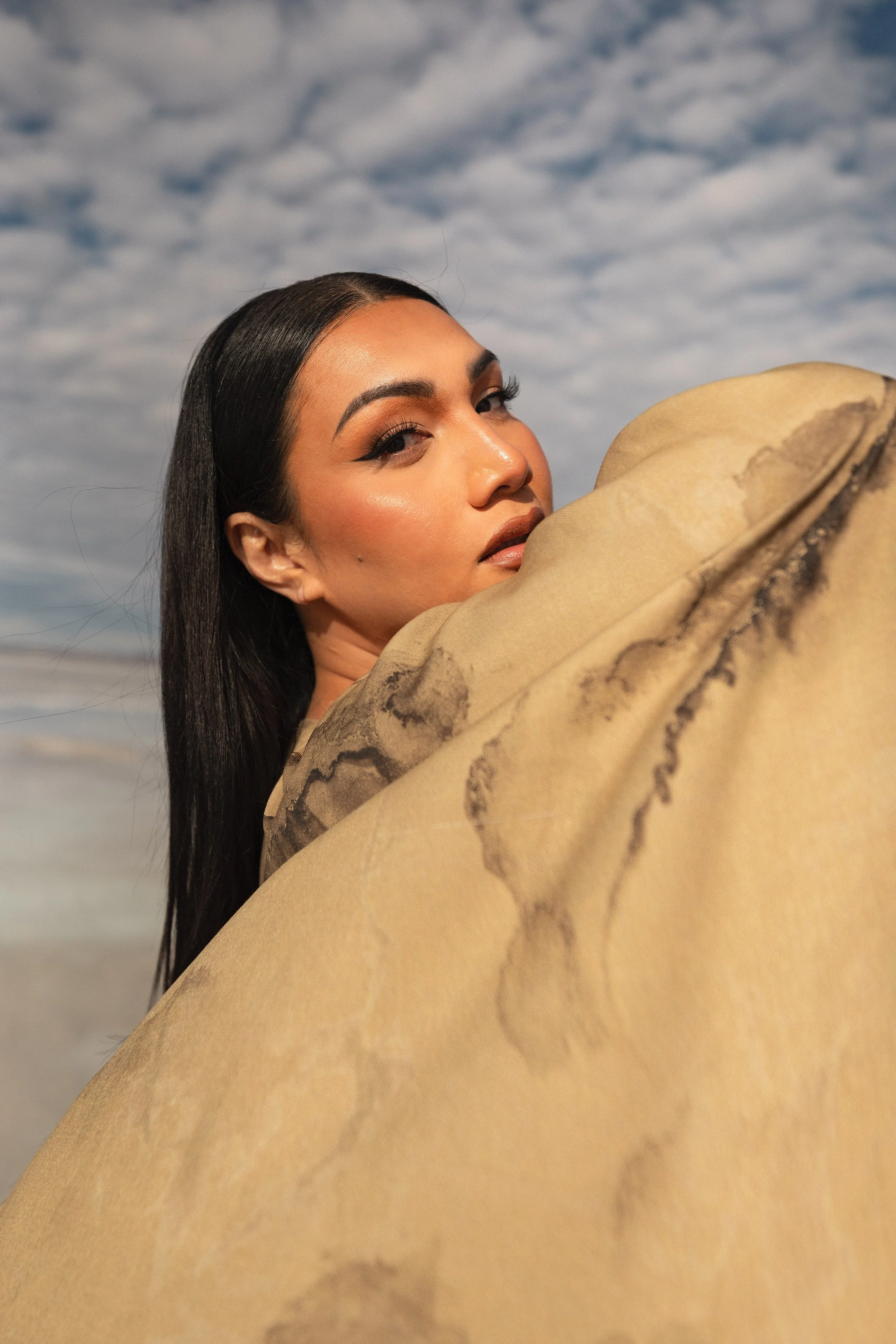 Close-up of a woman with long black hair, makeup, and tan clothing, looking over her shoulder against a cloudy sky background.