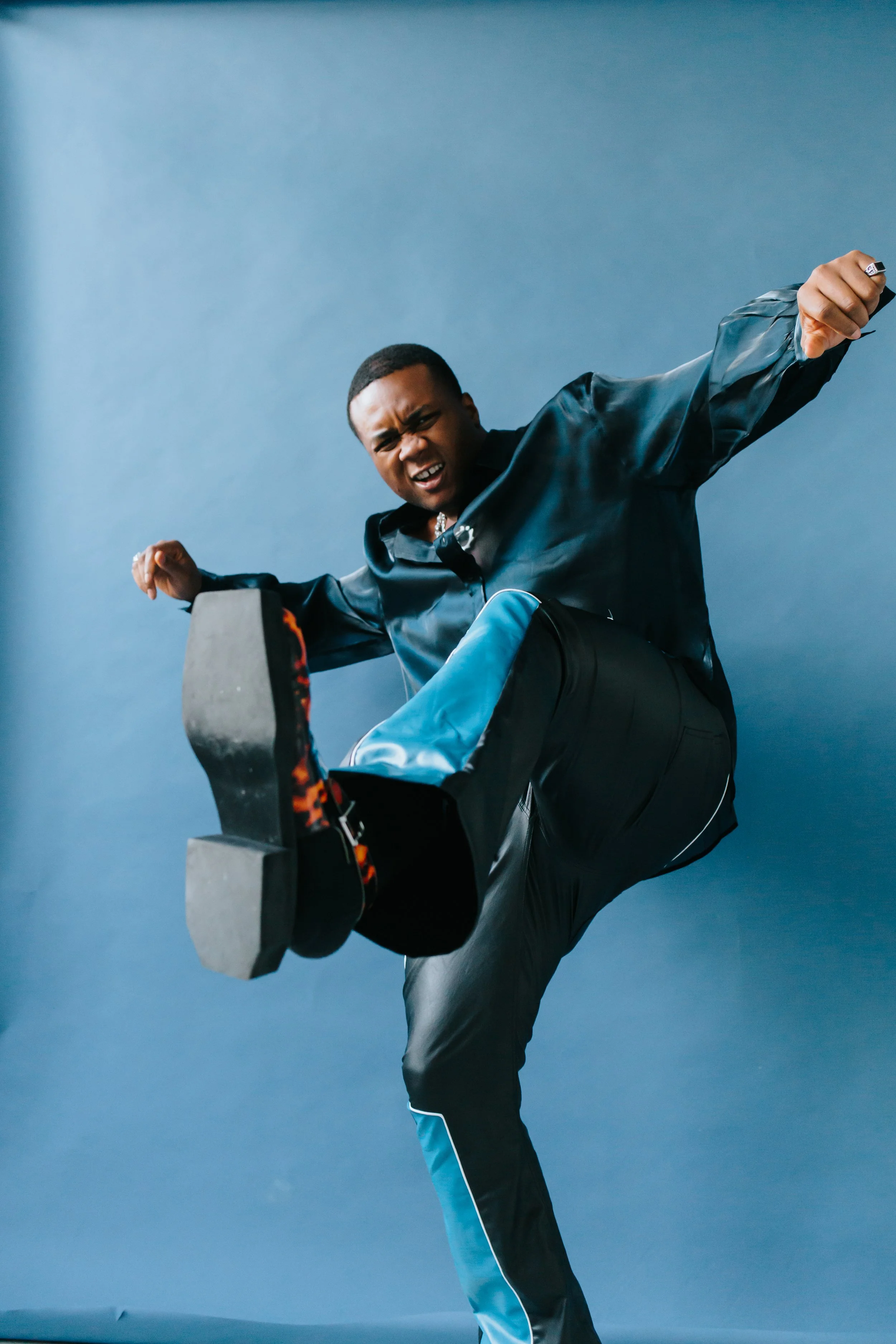 Young man in stylish black and blue outfit kicking with high energy against a blue background.