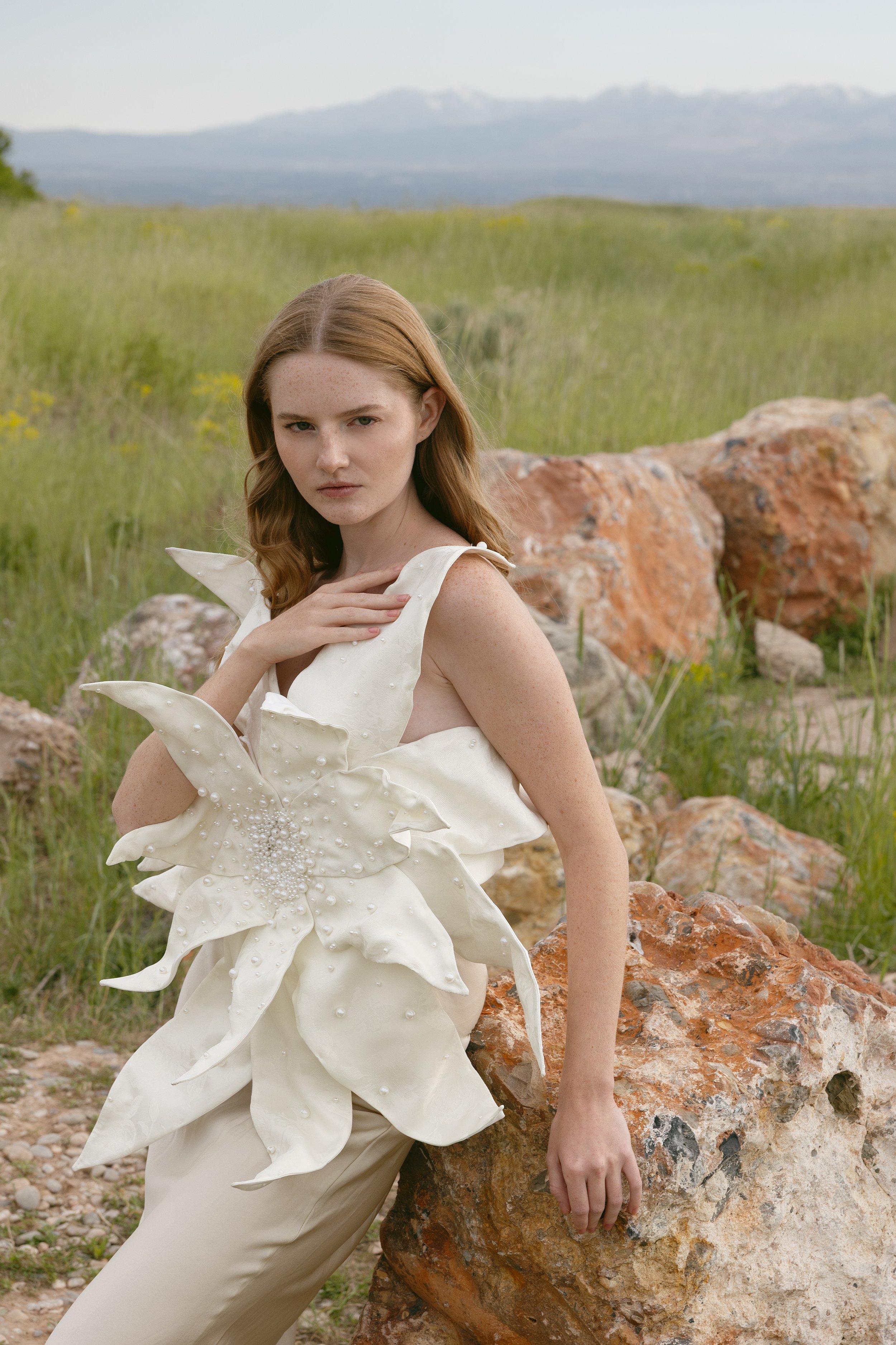 A young woman with long red hair stands outdoors in a grassy field with rocks, wearing an elaborate cream-colored dress with a large floral design. She has a serious expression and is touching her neck.