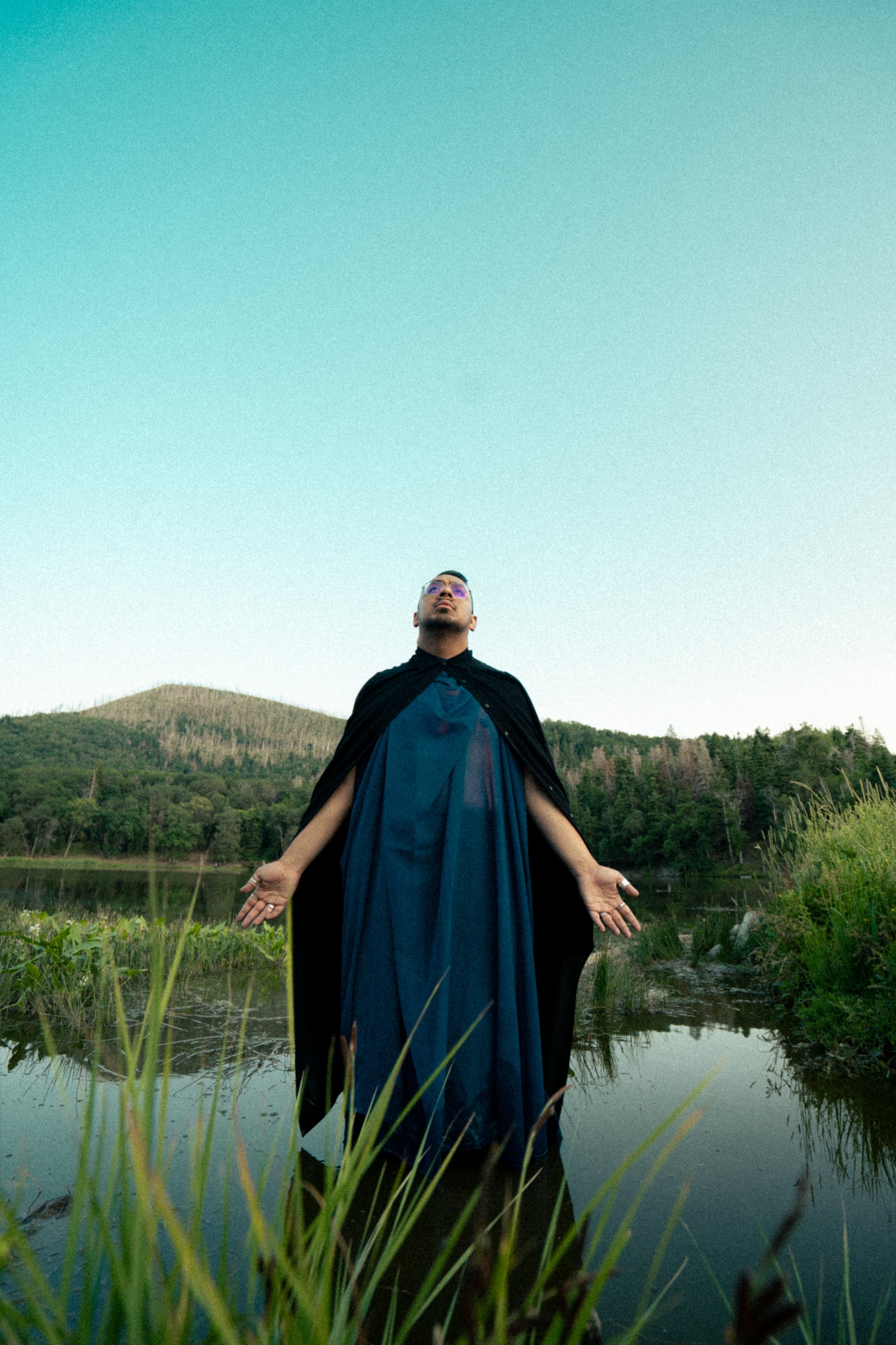 A person standing in a body of water with arms outstretched, wearing a dark cloak and a flowing blue dress, surrounded by nature with trees and hills in the background.