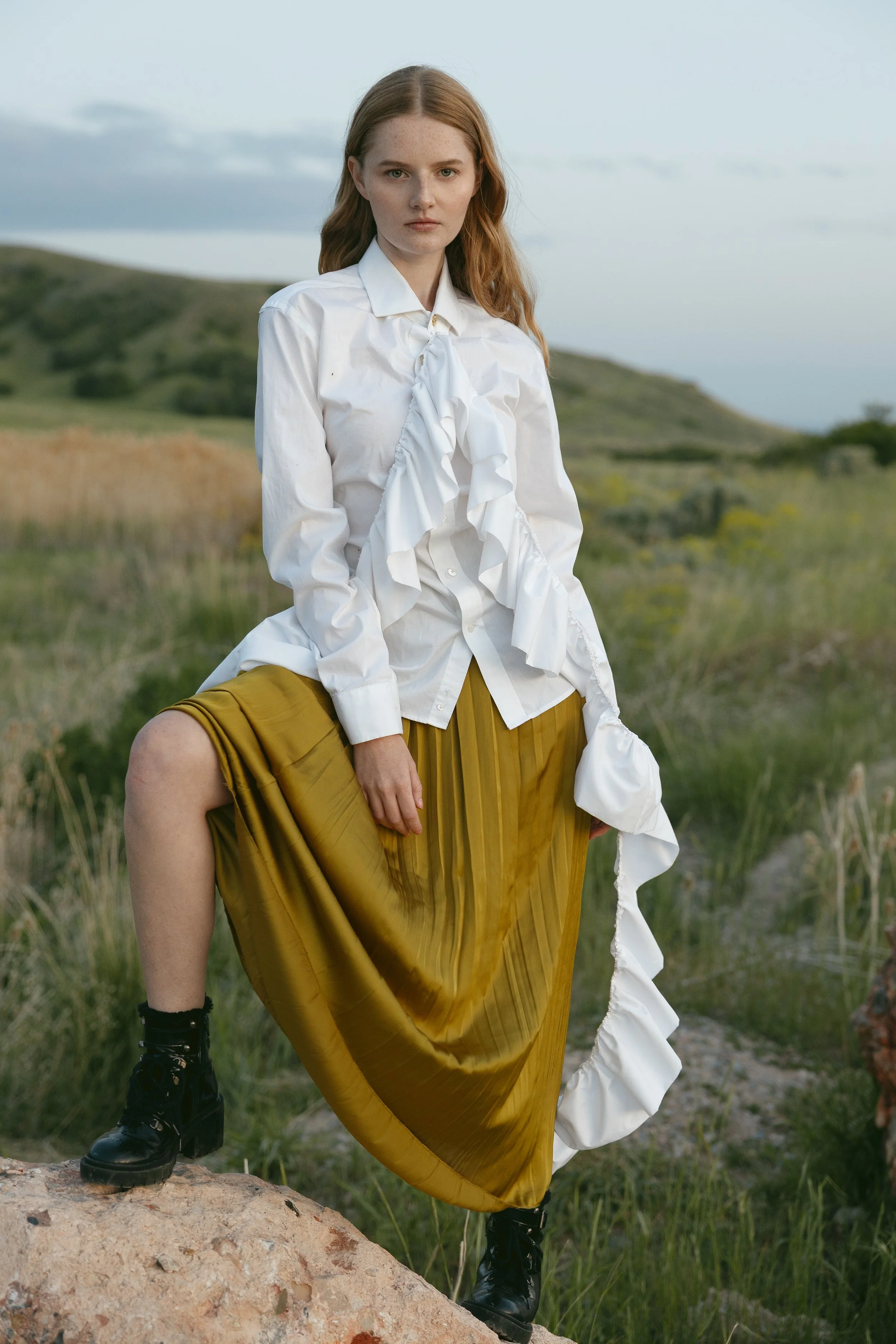 A young woman with long red hair standing outdoors on a rock in a grassy field with rolling hills in the background, wearing a white ruffled shirt, a mustard yellow pleated skirt, and black boots.