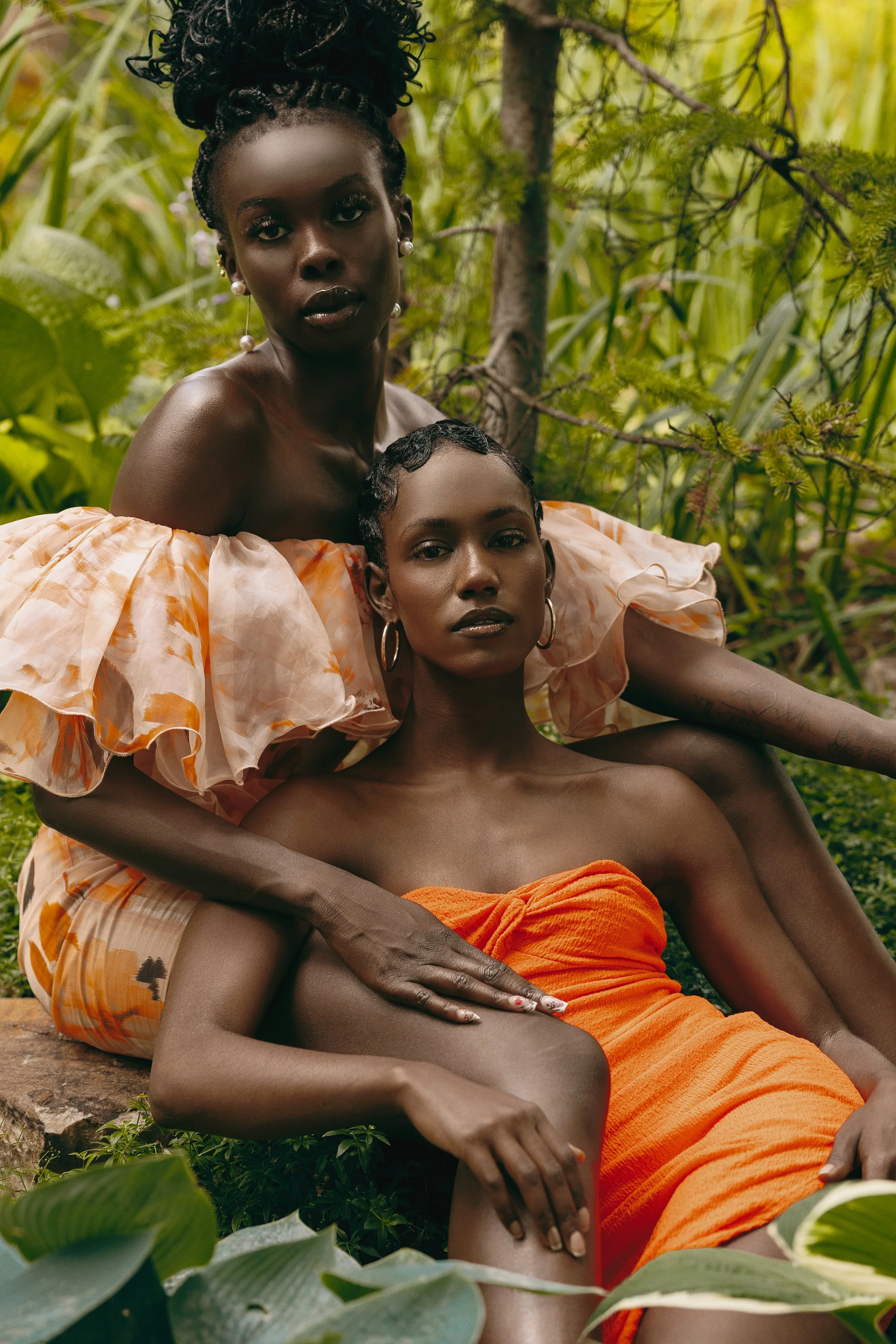 Two women with dark skin sitting outdoors in a lush green setting, both wearing elegant dresses. One woman is standing behind the other, gently resting her hand on her shoulder. They both gaze directly at the camera.
