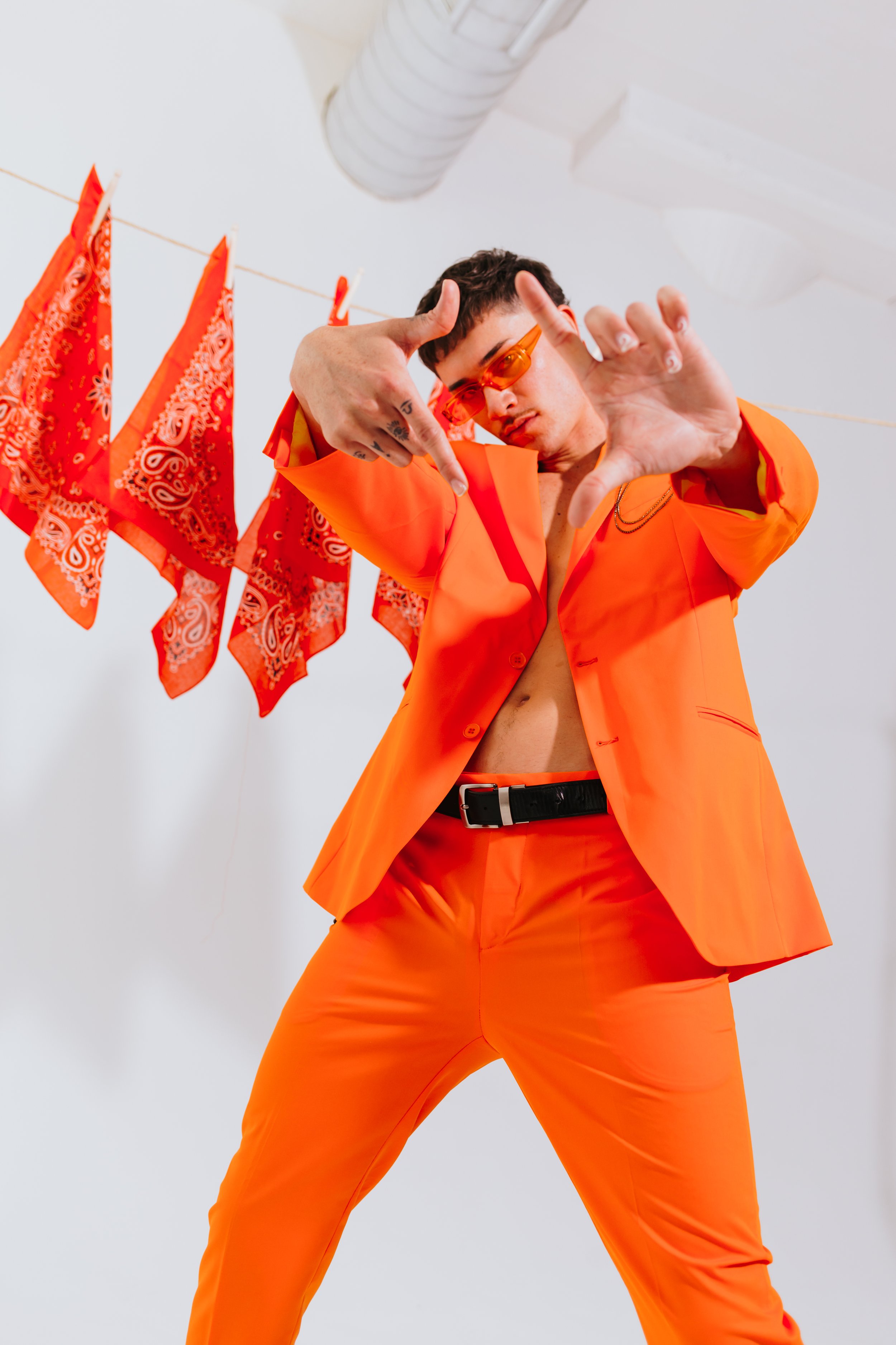 A person dressed in an orange suit and sunglasses poses with hands near their face, standing against a plain background with red bandanas hanging on a line.