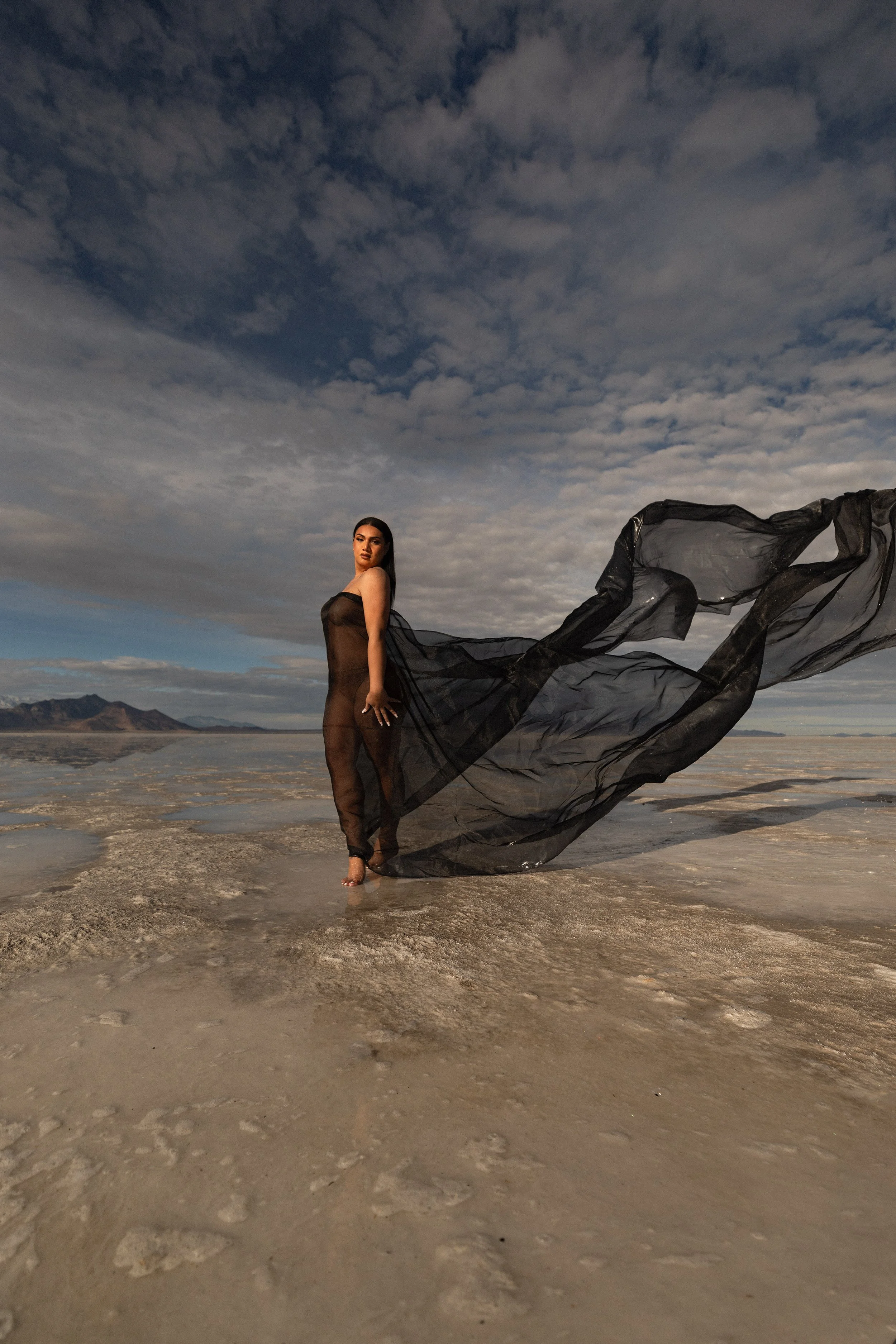 A woman in a sheer black dress stands barefoot on a salt flat with a flowing black fabric extending behind her. The sky is partly cloudy with distant mountains visible in the background.