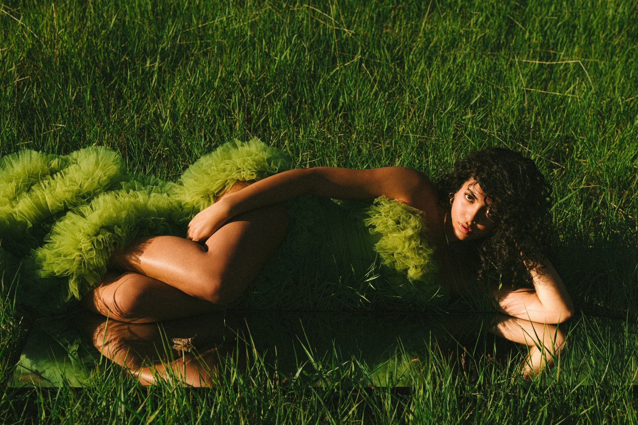 A woman in a green ruffled dress lying on her side in a grassy field, looking at the camera.