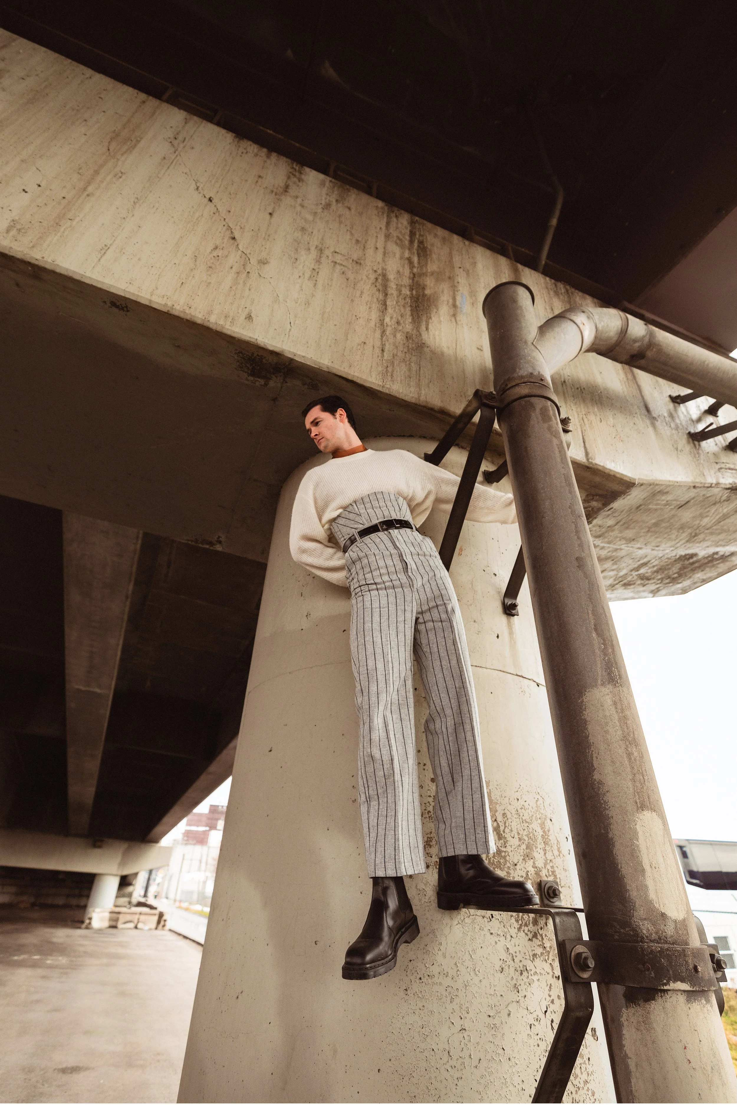 A woman dressed in a white sweater, gray plaid pants, and black boots is standing on a metal ladder under a concrete bridge or overpass.