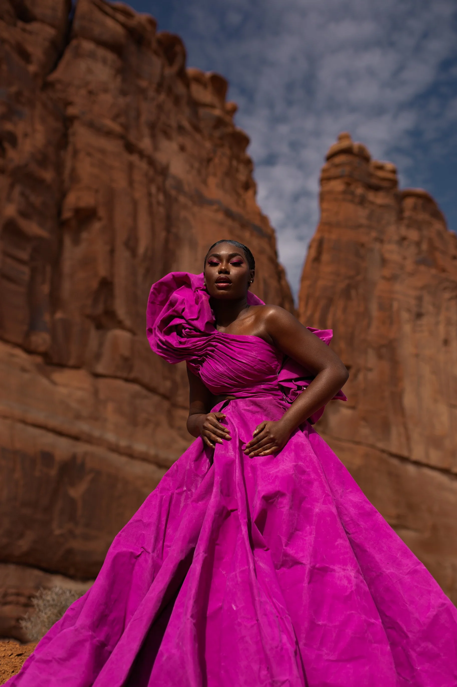A woman in a vibrant pink gown with dramatic ruffles and puffed sleeves, standing amidst red rock formations under a cloudy sky.