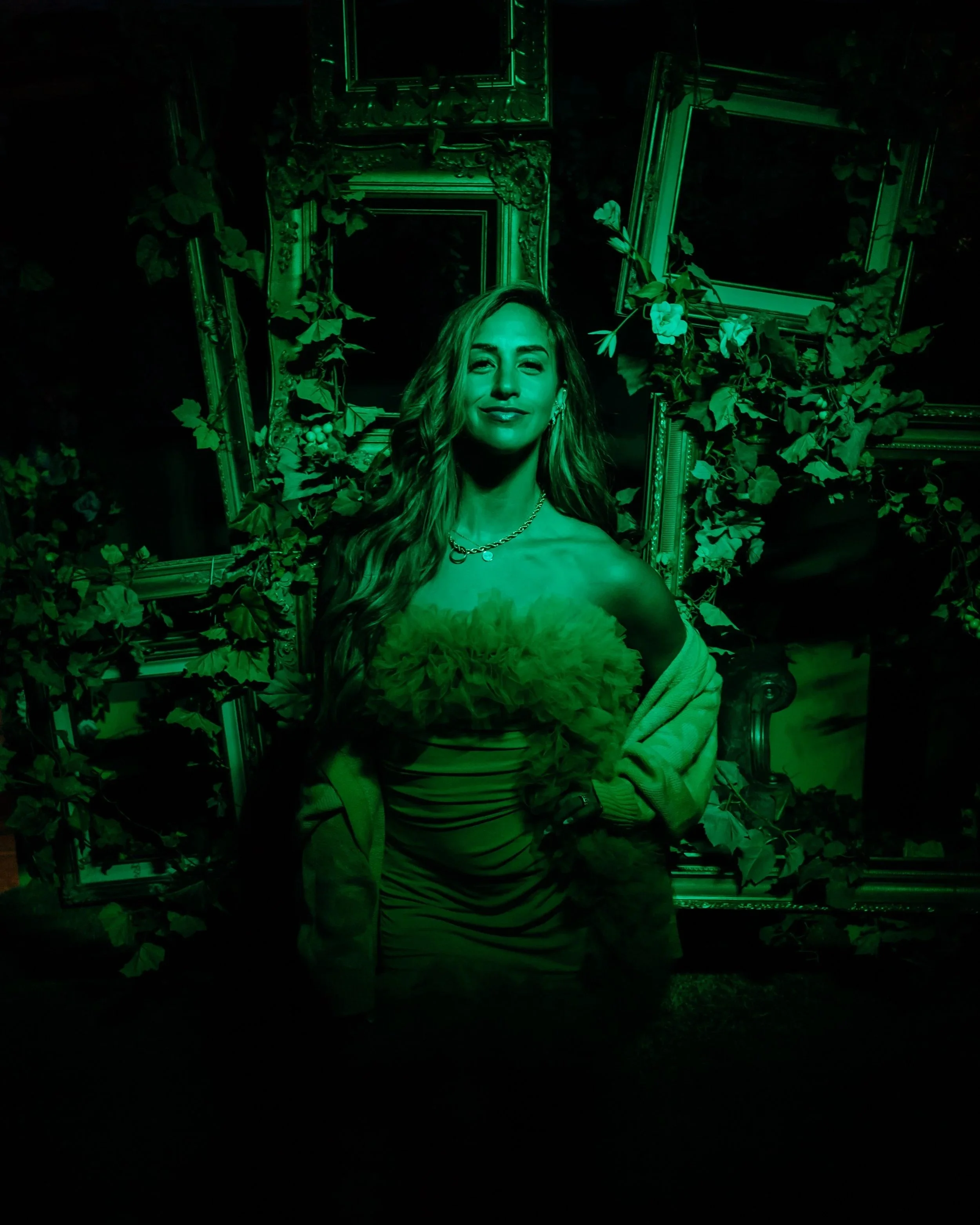A woman with long hair smiling, illuminated in green light, with empty picture frames and green foliage behind her.