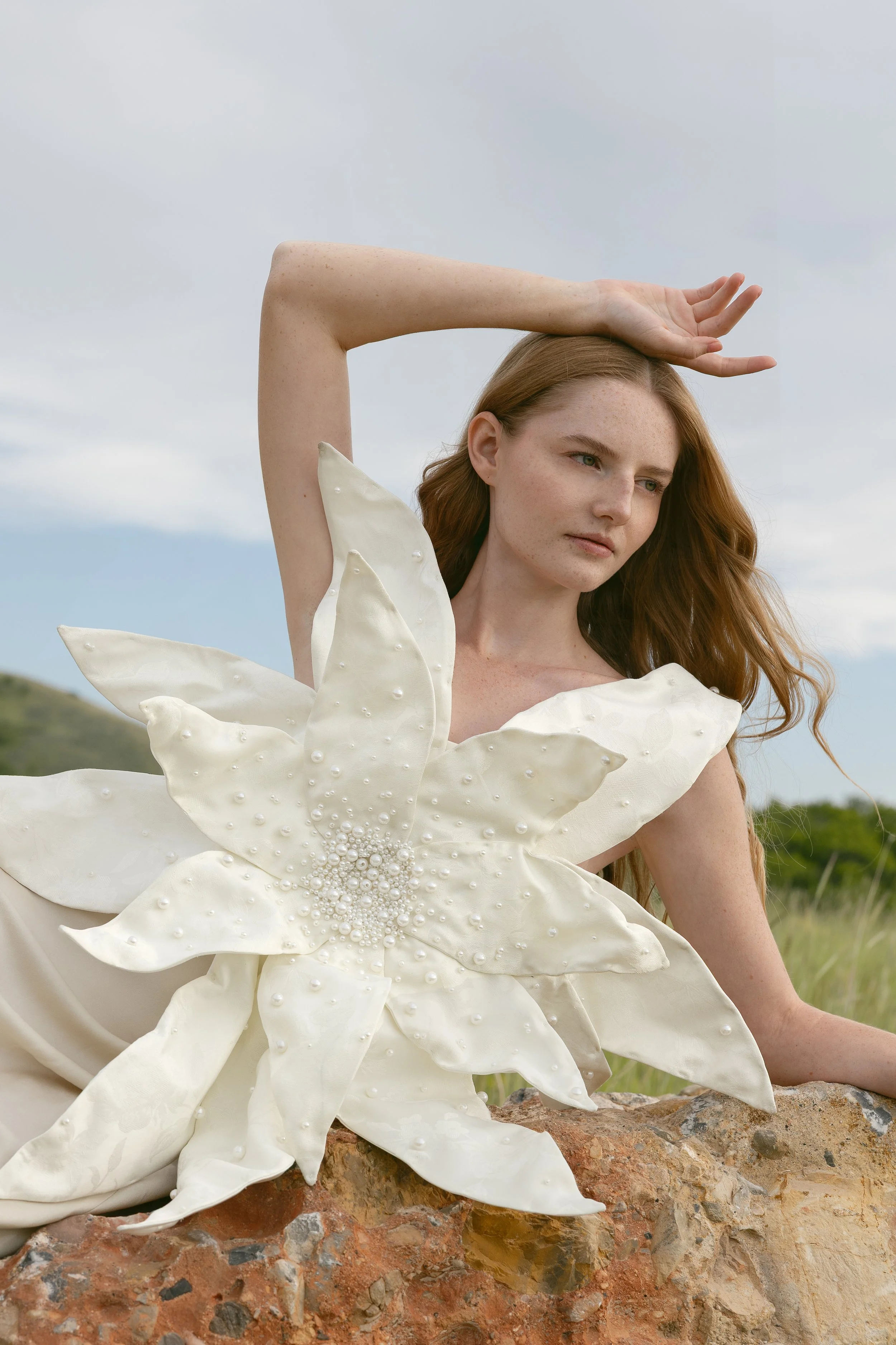 A woman with long red hair outdoors, dressed in a cream-colored gown with large floral adornment on her chest, sitting on a rock with a cloudy sky and distant greenery in the background.